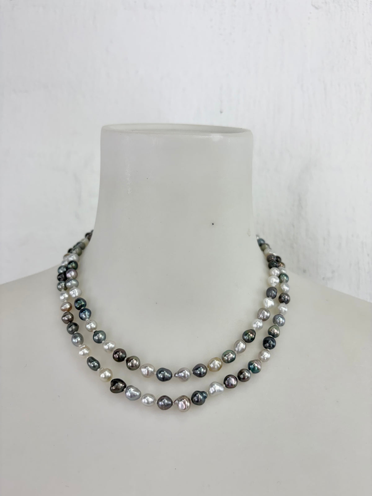 A double-strand necklace of irregular baroque pearls in shades of gray, black, and white, displayed on a white mannequin bust against a white textured wall.