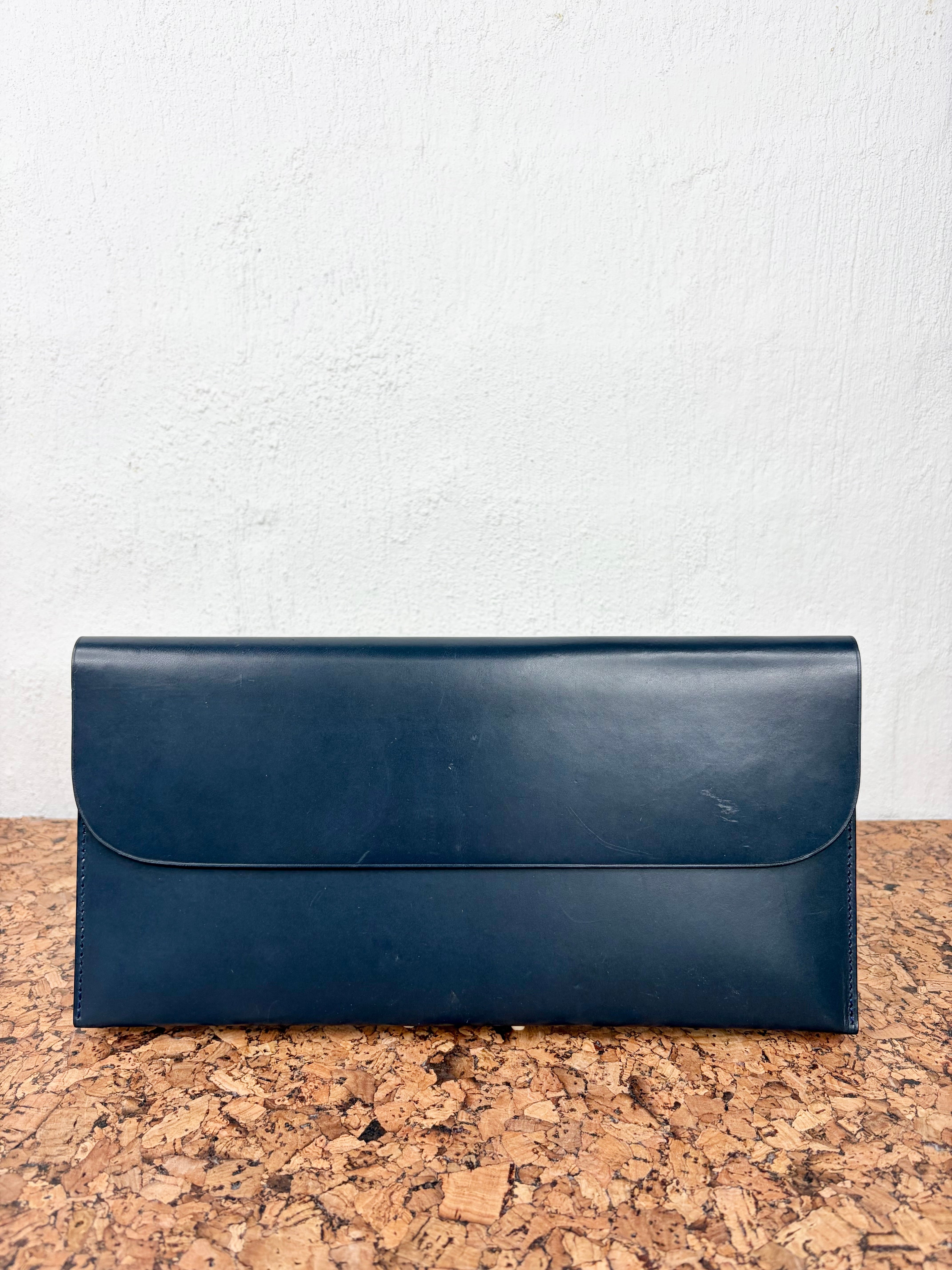 A dark navy blue leather clutch or envelope-style wallet with a flap closure, resting on a cork surface against a white textured wall.