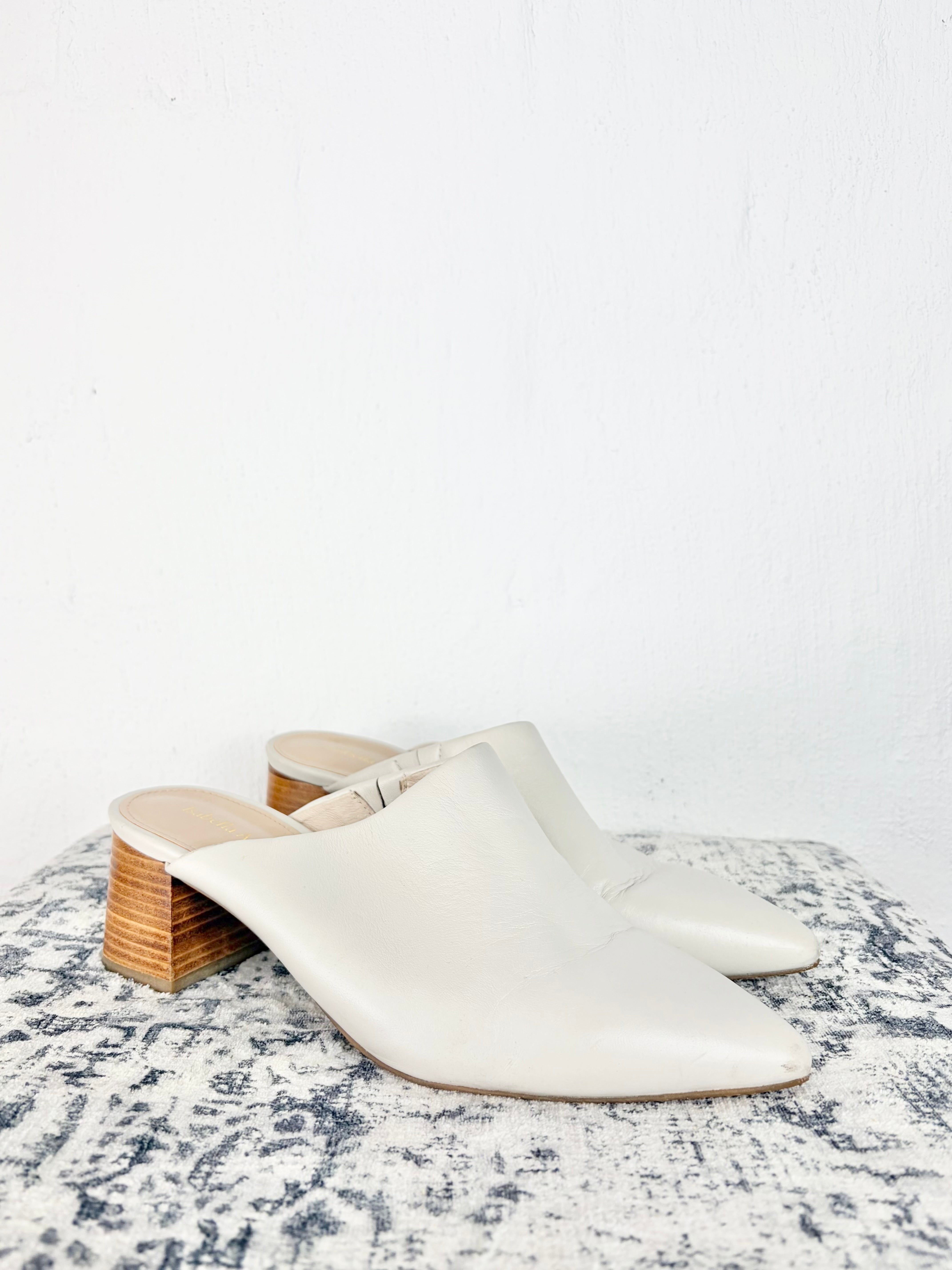 A pair of off-white leather mule heels with chunky stacked wood block heels, displayed on a distressed gray and white patterned surface against a white wall.