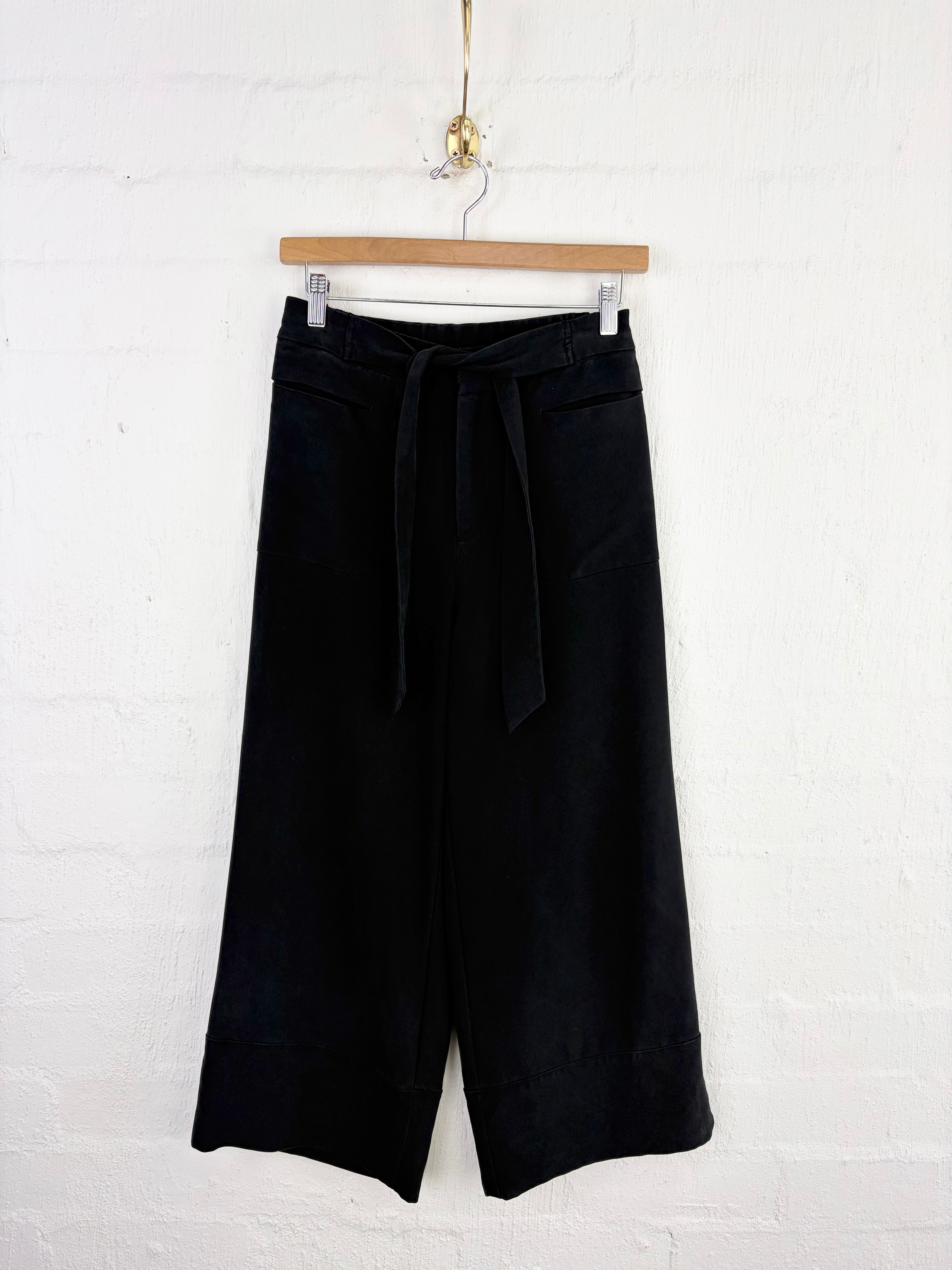 Black wide-leg cropped pants with a matching fabric tie belt, hanging on a wooden hanger against a white painted brick wall.