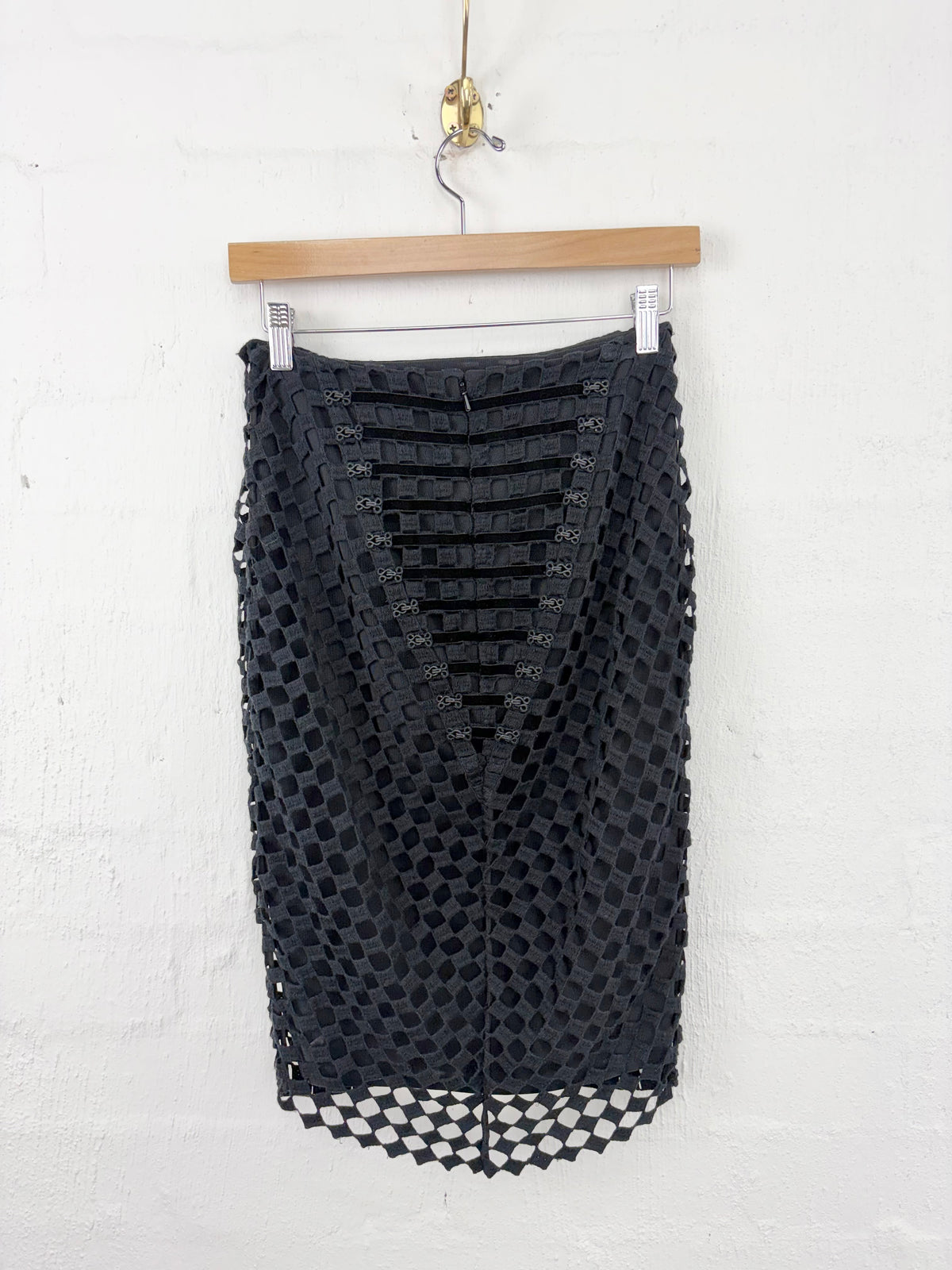 A black crochet knit skirt with a V-neckline and side cutouts, hanging from a wooden hanger against a white textured wall.