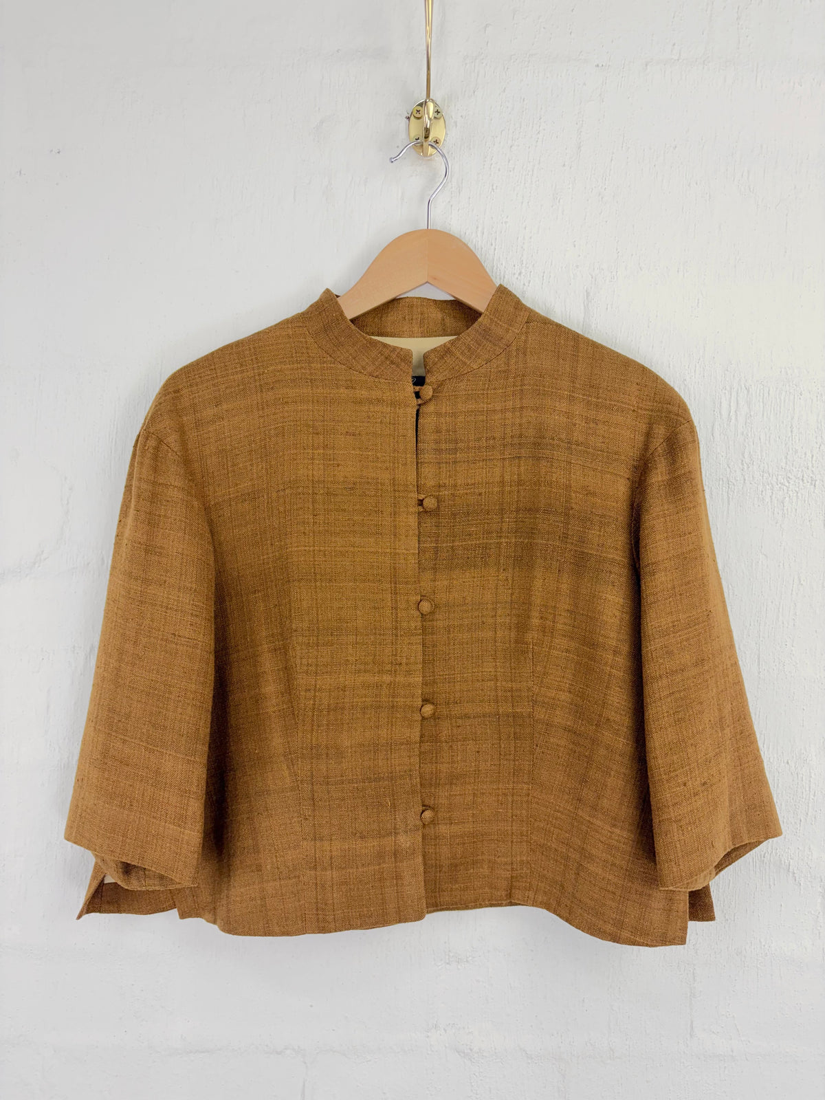 A textured mustard-brown linen jacket with a mandarin collar and button front, hanging on a wooden hanger against a white brick wall.