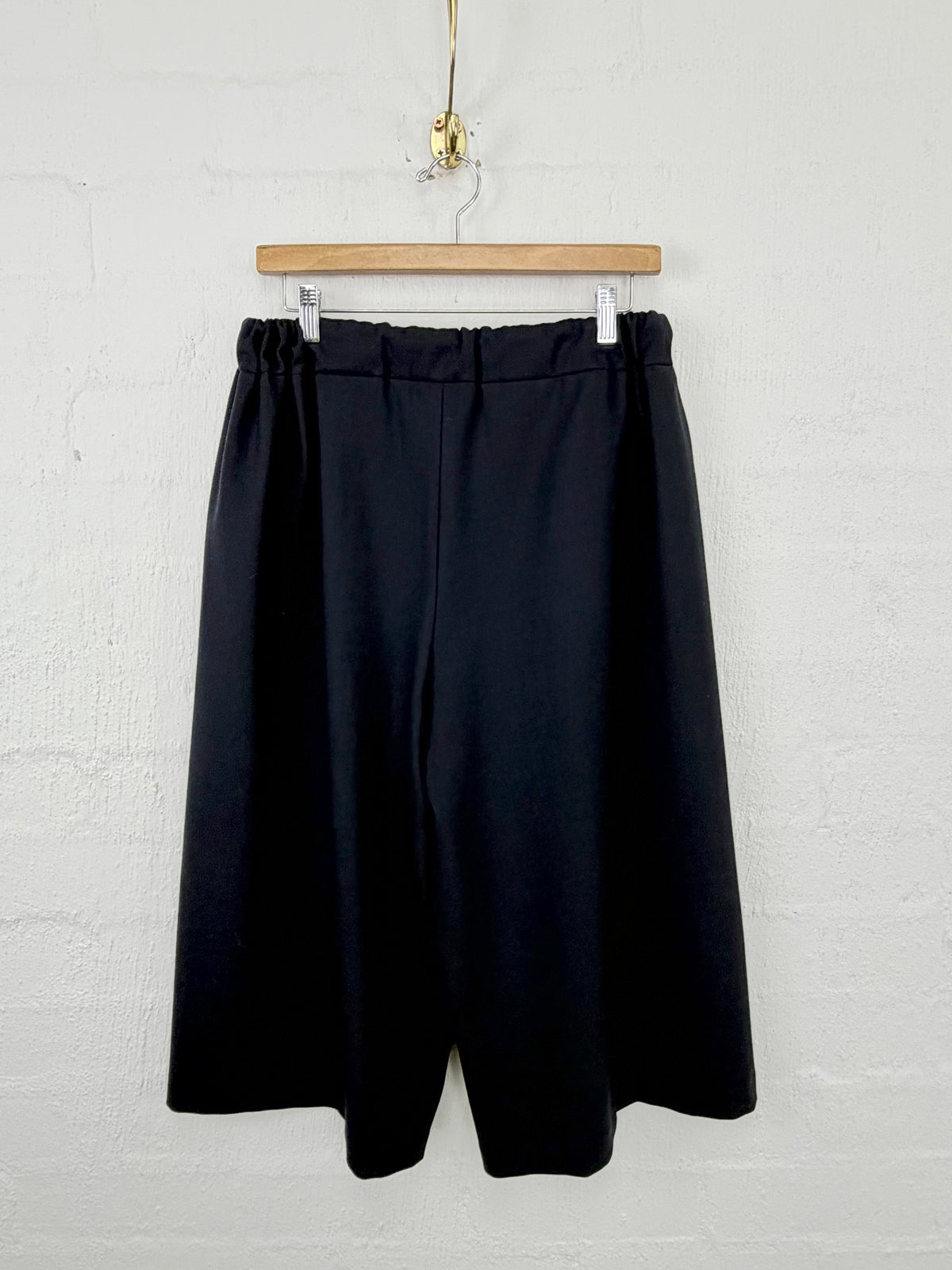Black wide-leg cropped trousers hanging from a wooden hanger against a white painted brick wall.