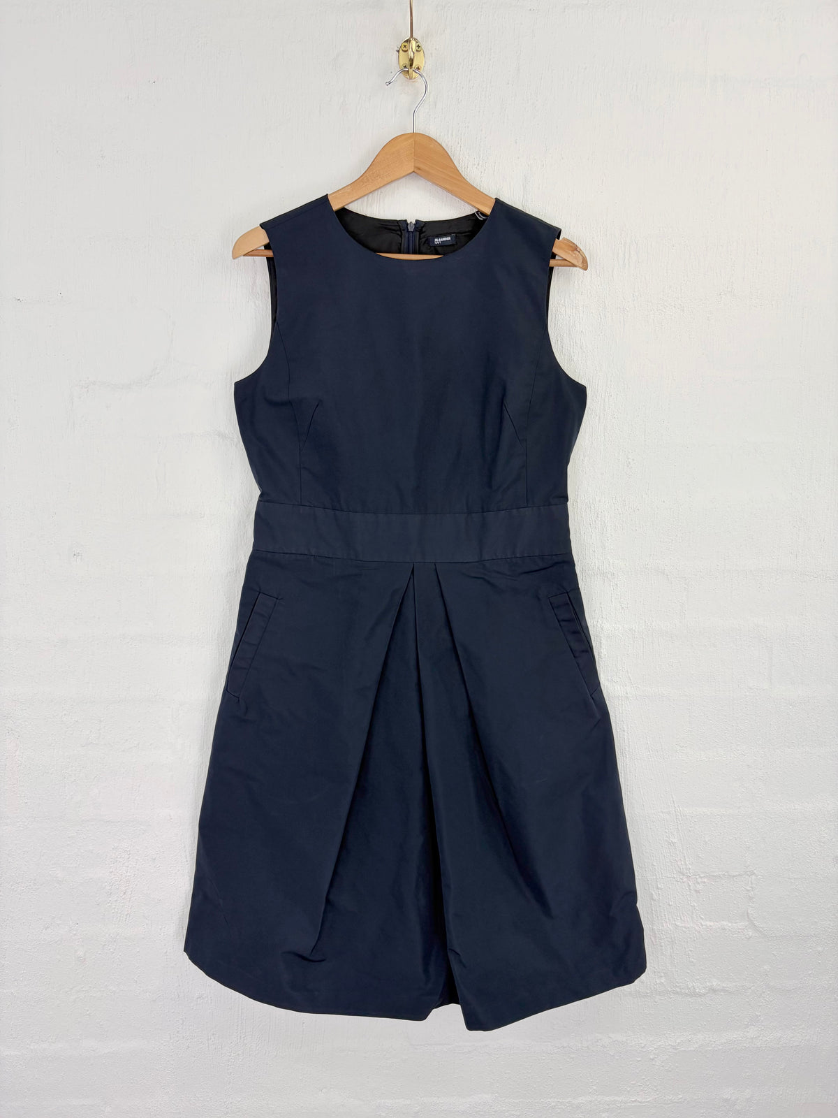 A sleeveless navy blue dress with a pleated skirt detail, hanging on a wooden hanger against a white painted brick wall.