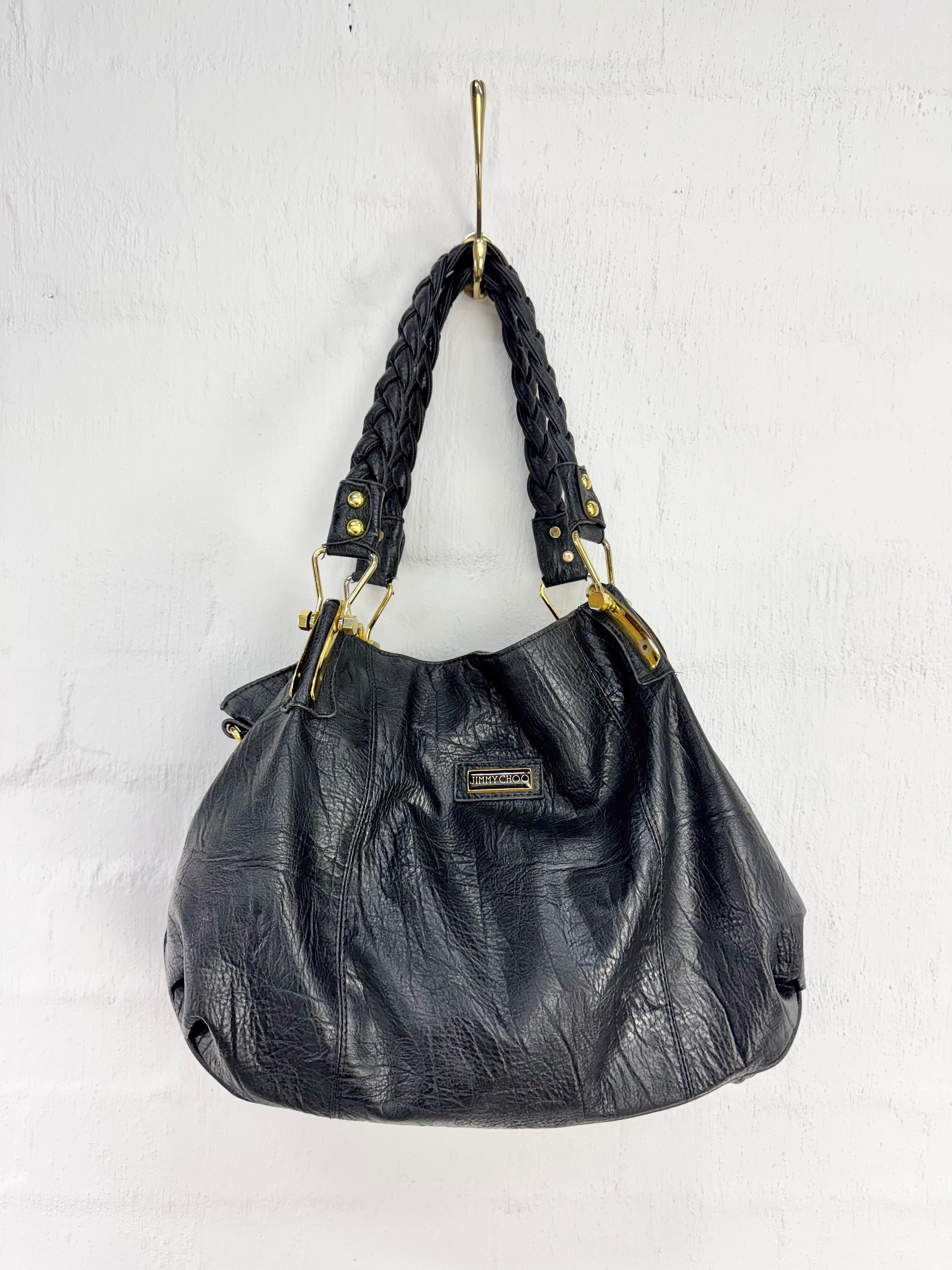 A black textured leather slouchy handbag with braided shoulder straps and gold-tone hardware, hanging from a gold hook against a white brick wall.