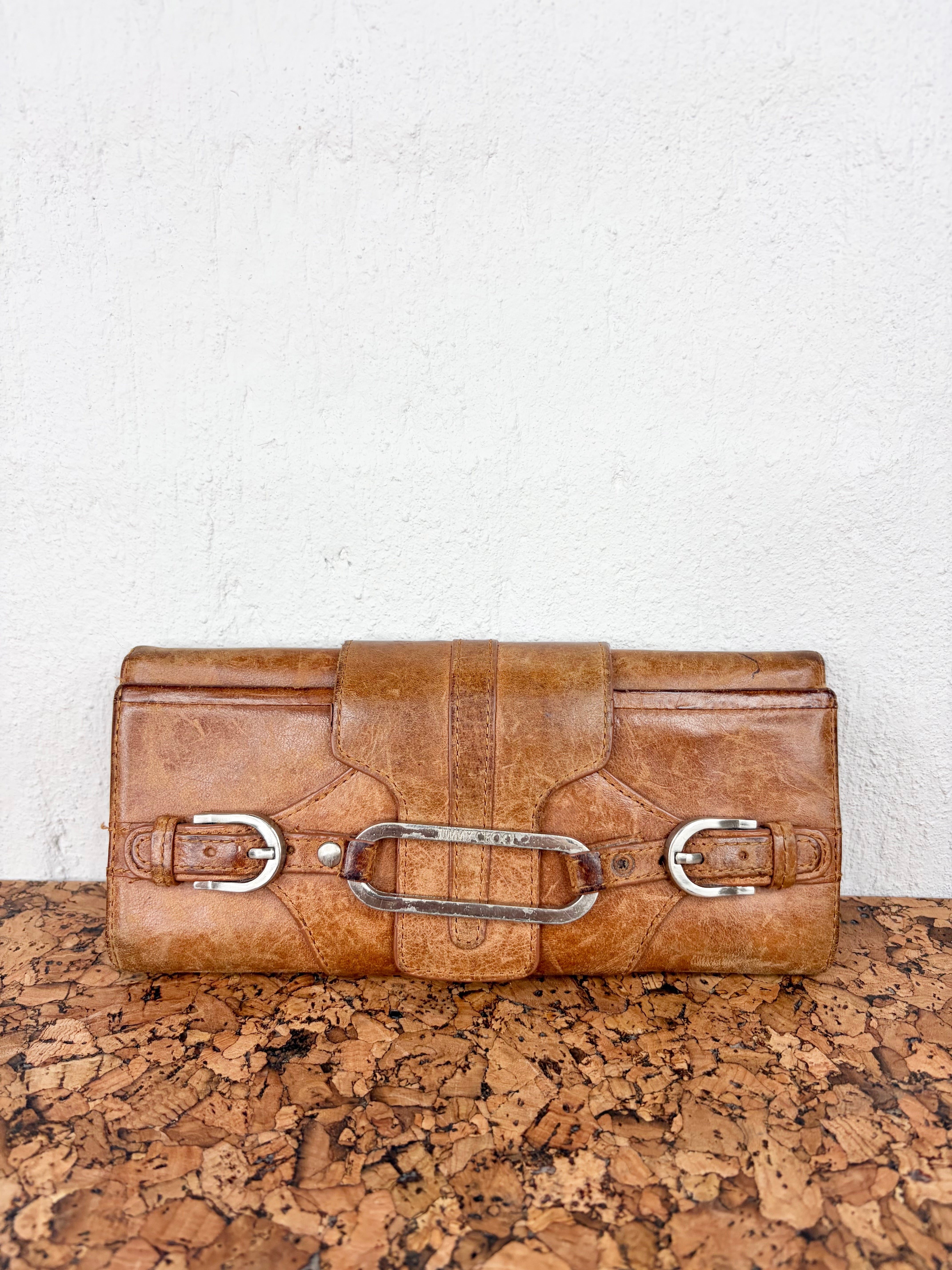 A distressed brown leather clutch bag with silver-tone hardware, resting on a textured cork surface against a white wall.