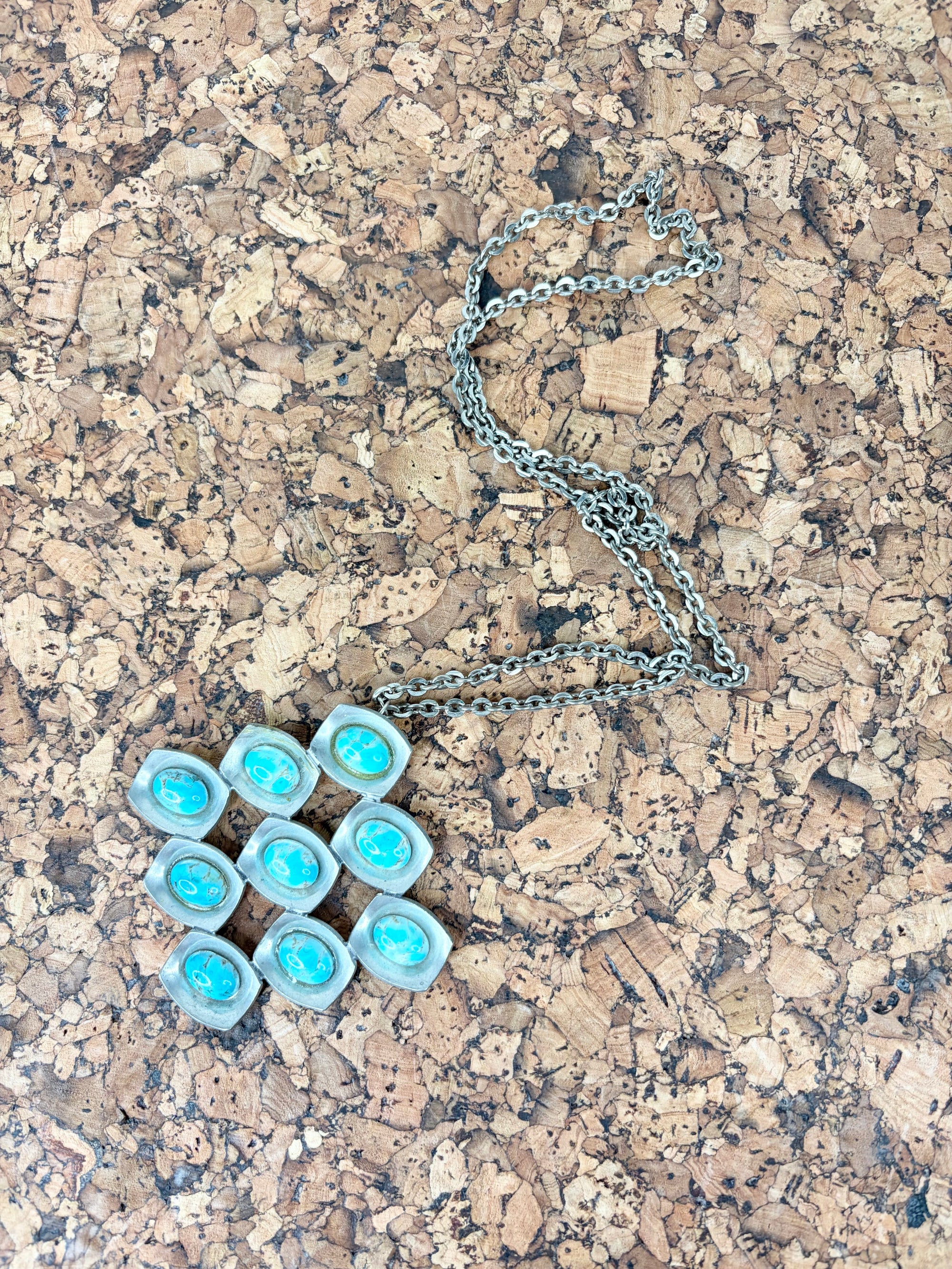 A silver chain necklace with a pendant made of nine small turquoise-blue oval stones set in silver, displayed on a cork surface.