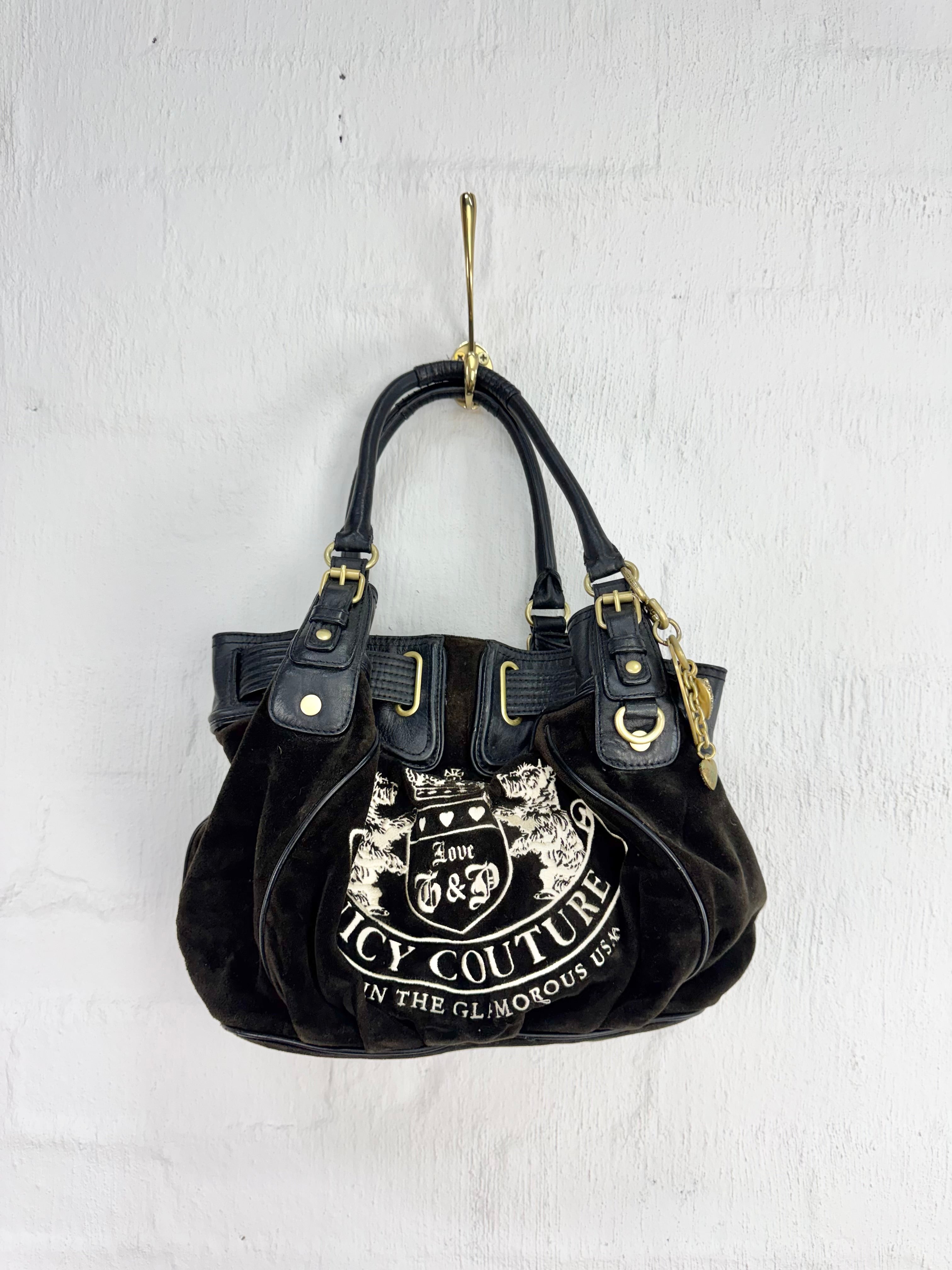 A black leather handbag with gold-tone hardware and a large white 'Juicy Couture' graphic on the front, hanging from a gold hook against a white textured wall.