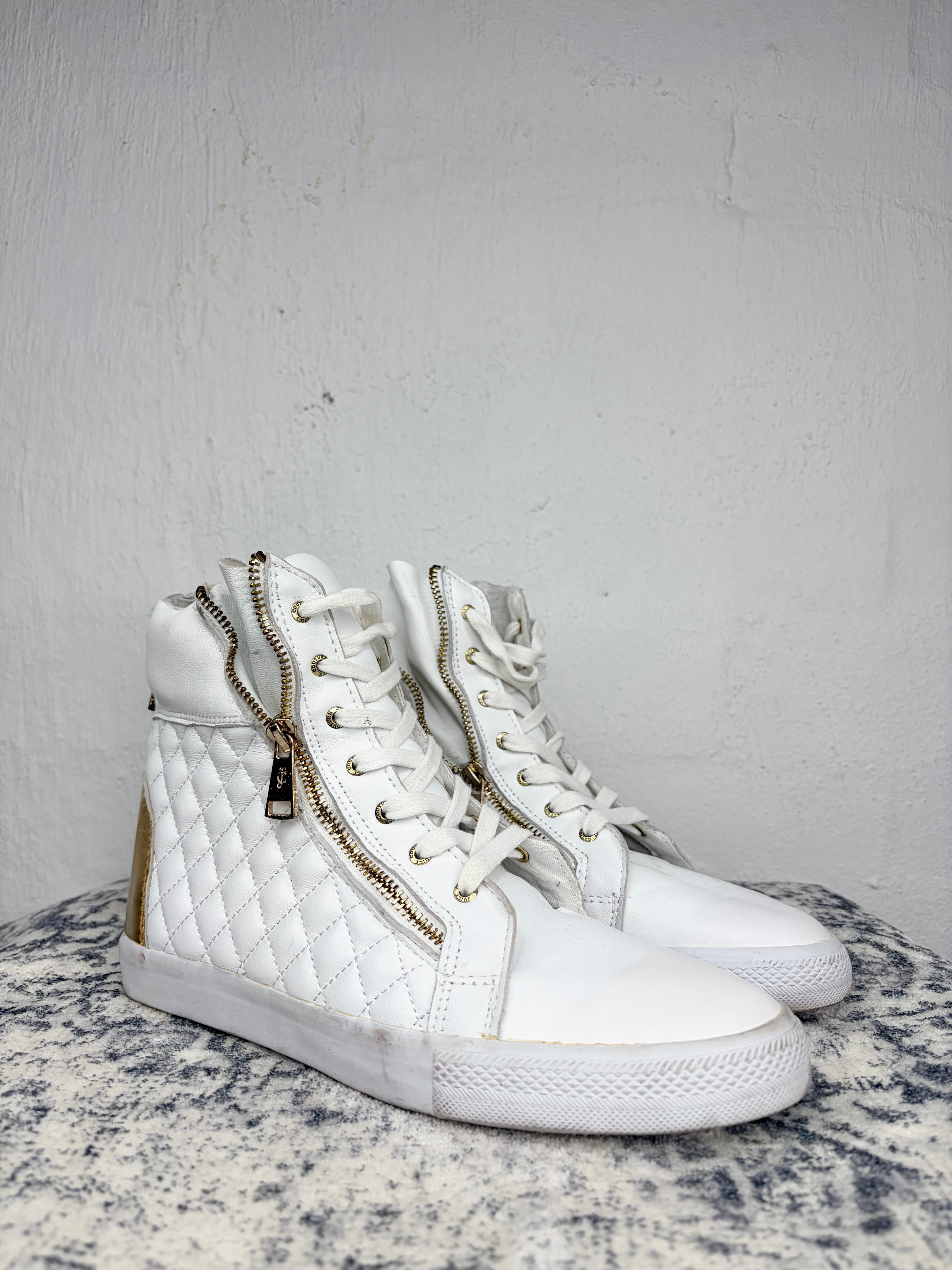 A pair of white high-top sneakers with quilted side panels, gold-tone zippers, and white laces, displayed on a speckled gray surface against a textured light gray wall.