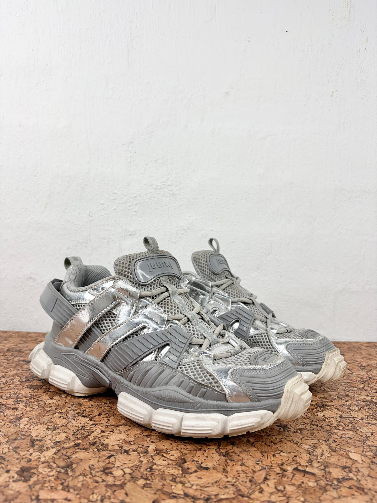 A pair of chunky, multi-layered athletic sneakers in metallic silver and gray, featuring mesh panels, synthetic overlays, and thick sculpted soles, resting on a brown cork surface against a white wall.