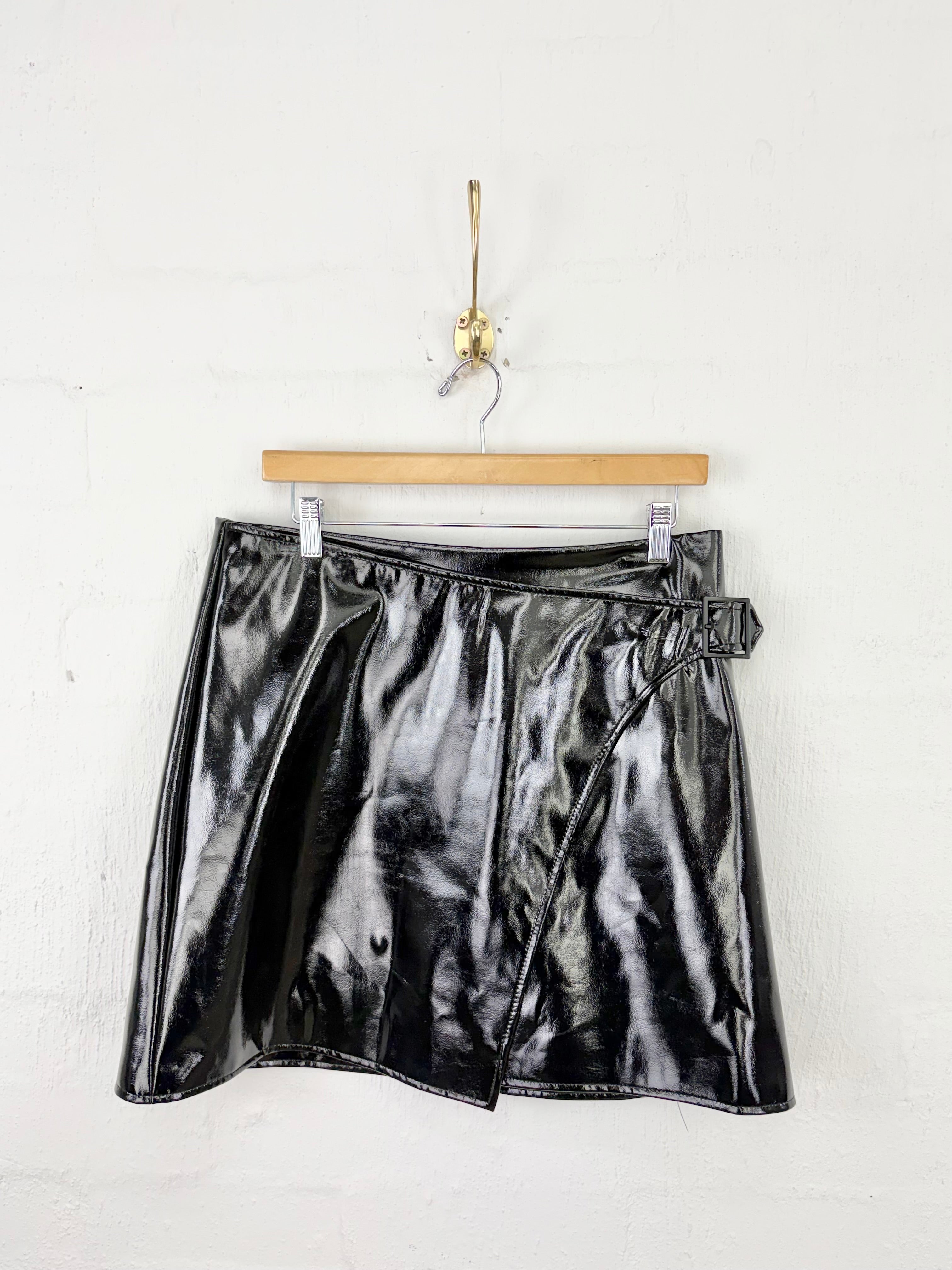 A shiny black faux-leather wrap mini skirt hanging from a wooden hanger on a white brick wall.