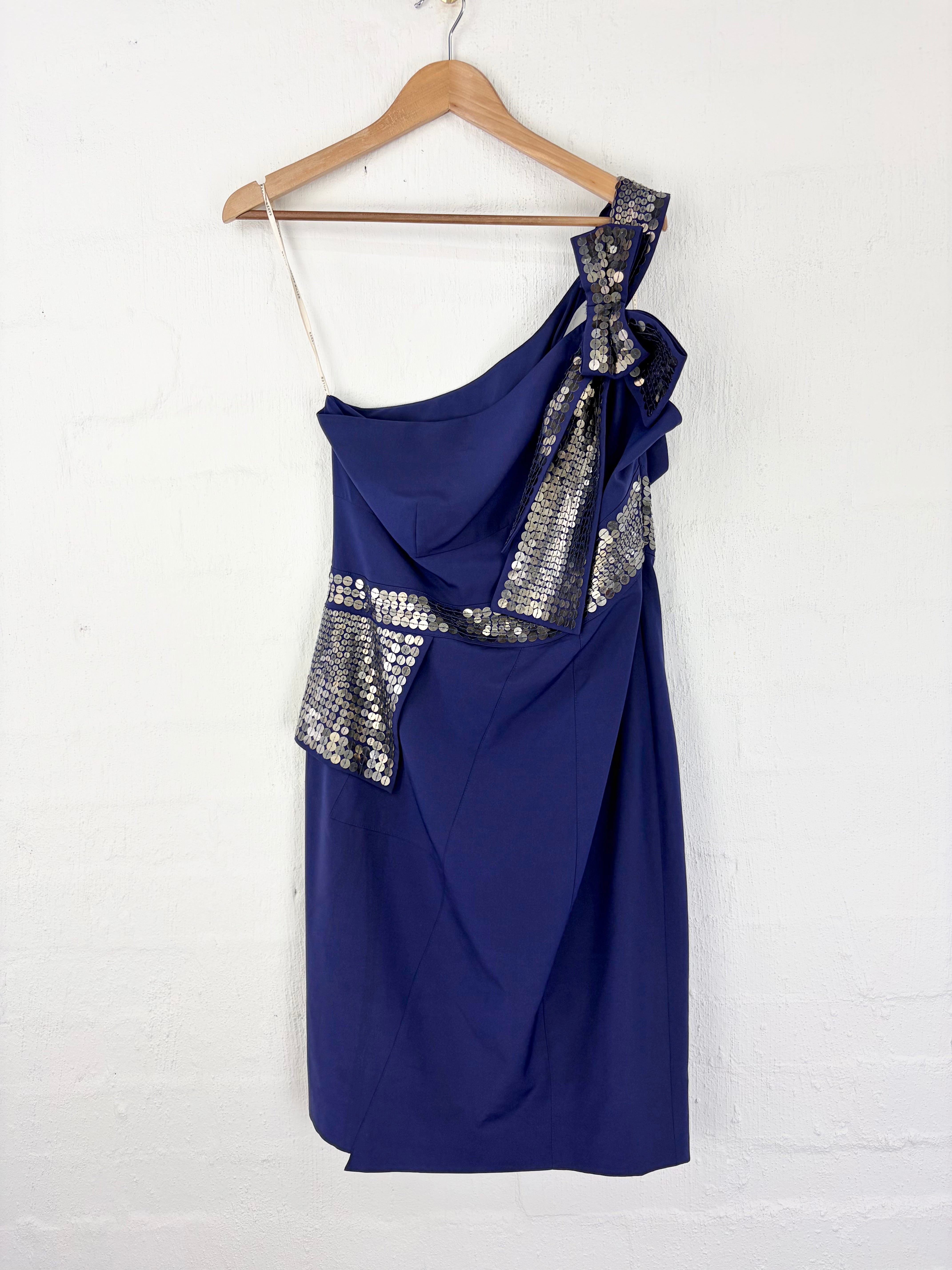 A navy blue one-shoulder dress with silver sequined ruffle accents, hanging on a wooden hanger against a white painted brick wall.