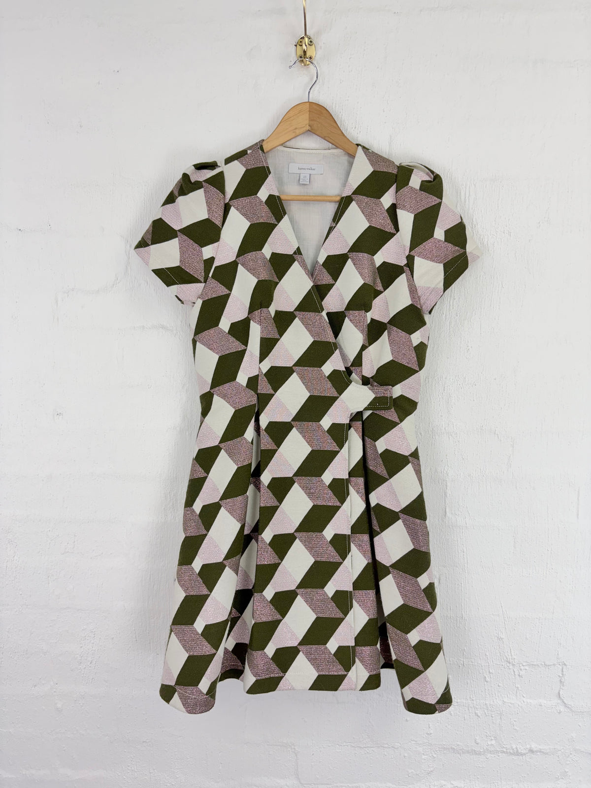 A short-sleeve wrap dress with a geometric 3D cube pattern in olive green, dusty pink, and cream, hanging on a wooden hanger against a white painted brick wall.