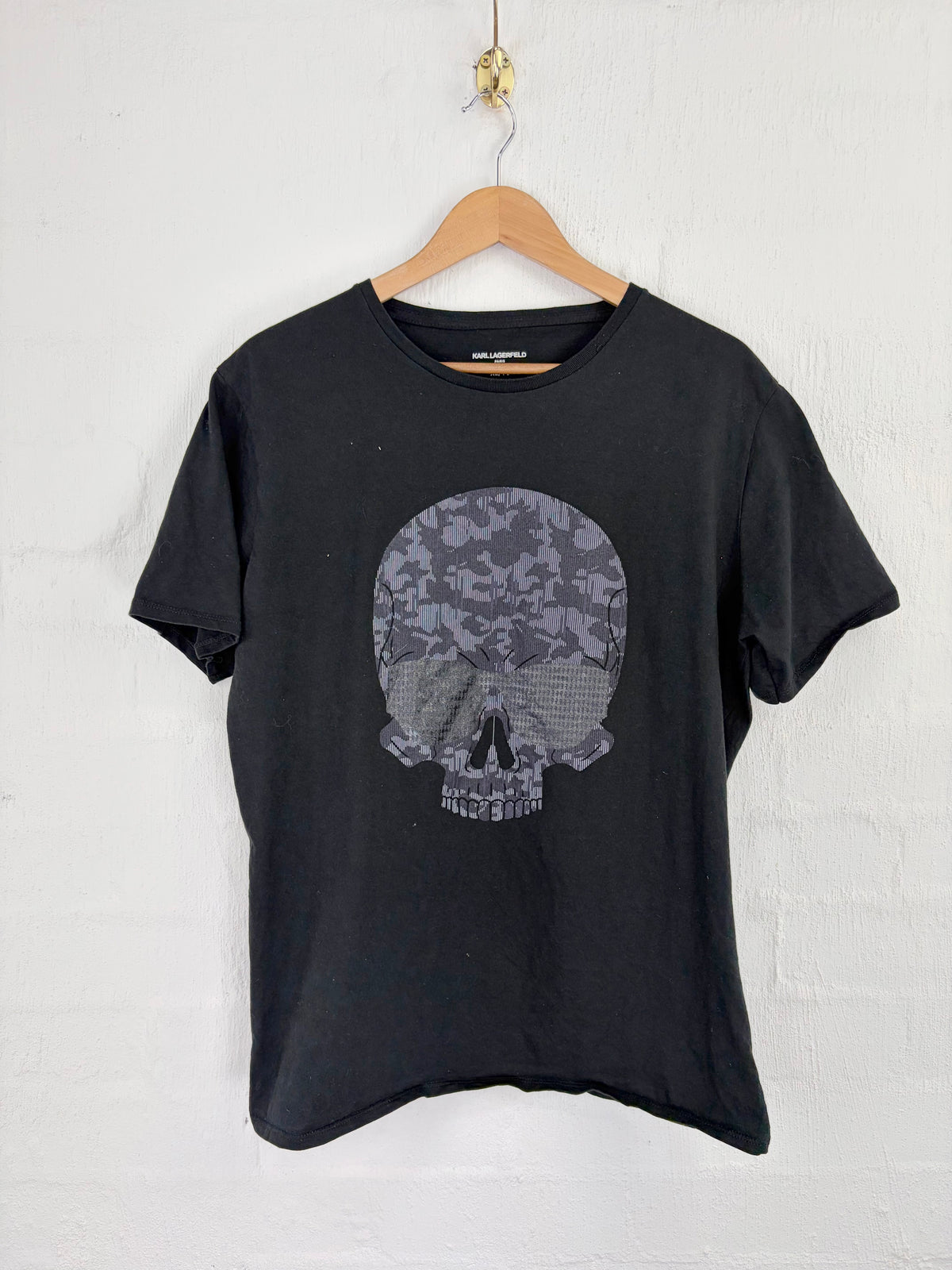 A black short-sleeve t-shirt hanging on a wooden hanger against a white painted brick wall. The shirt features a large graphic print of a skull on the front, filled with a gray and black floral pattern.