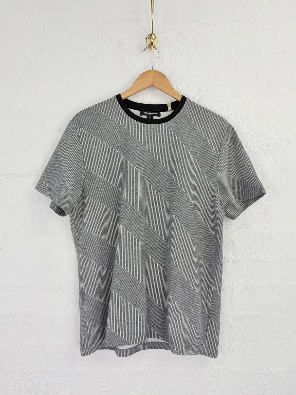 A gray short-sleeve knit t-shirt with a textured diagonal stripe pattern and a black crew neck, hanging on a wooden hanger against a white brick wall.
