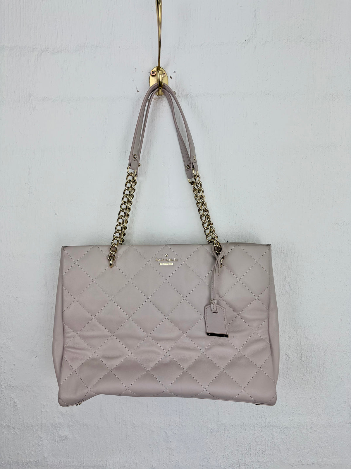 A light pink quilted leather tote bag with gold-tone chain shoulder straps, hanging from a gold hook against a white painted brick wall.