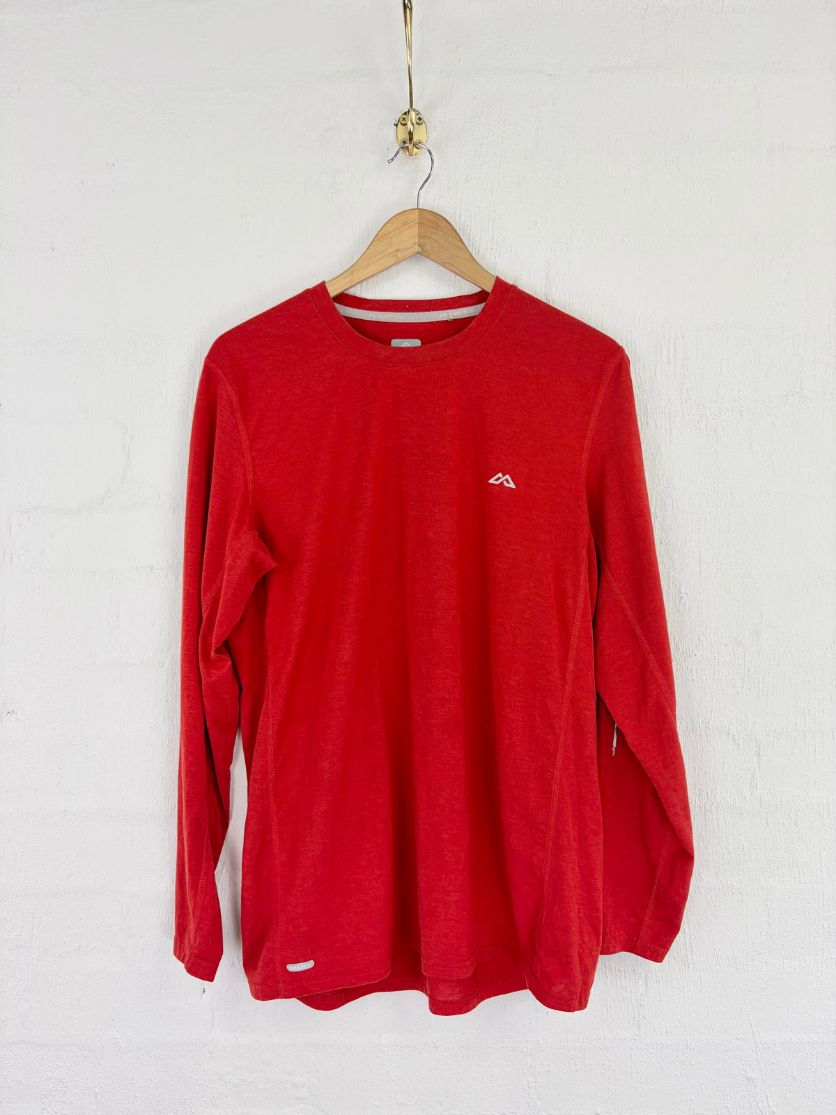 A red long-sleeve crewneck shirt hanging on a wooden hanger against a white textured wall. A small white mountain logo is visible on the left chest.