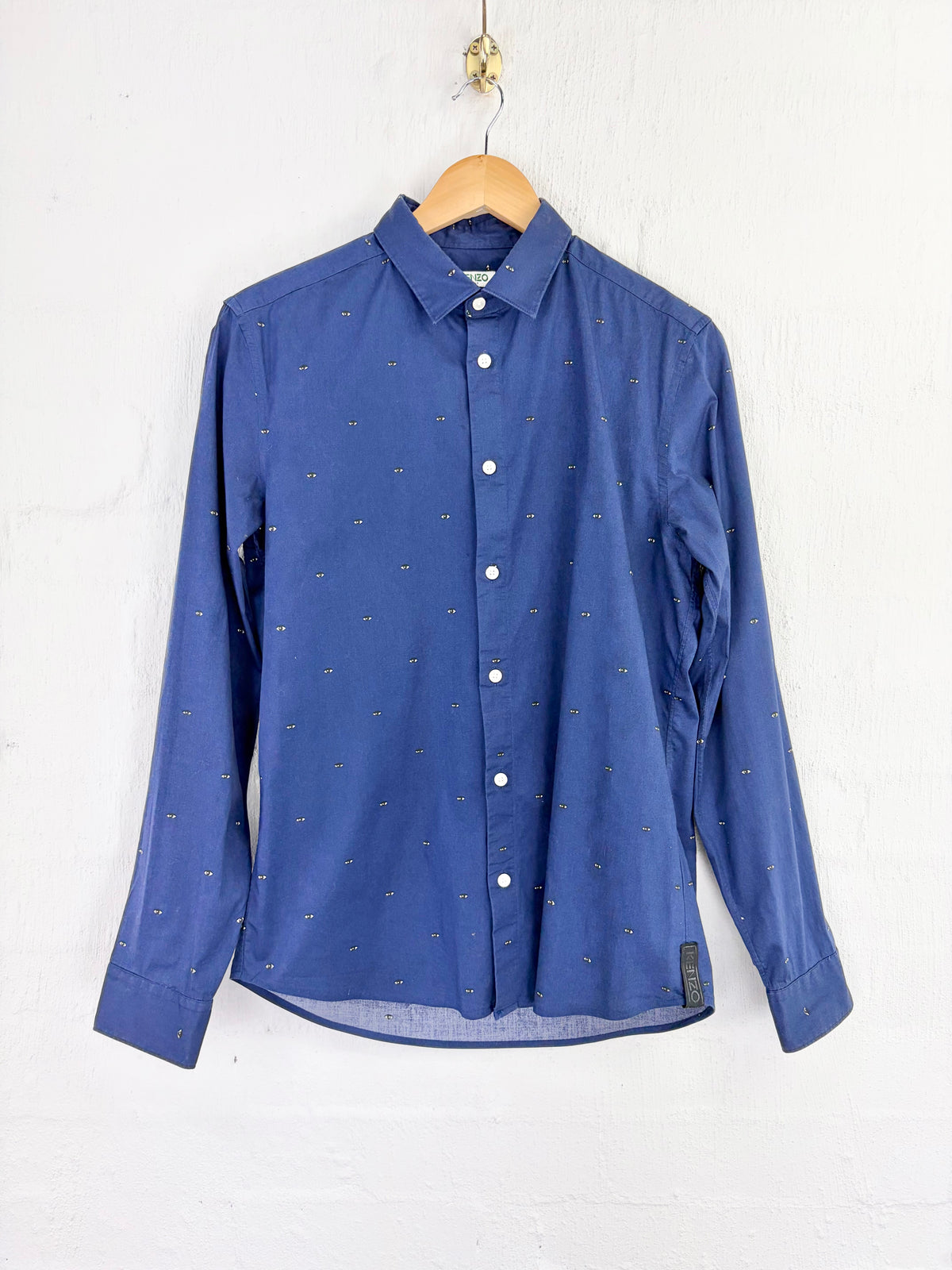 A blue long-sleeve button-up shirt with a tiny repeating cross pattern, hanging on a wooden hanger against a white painted brick wall.