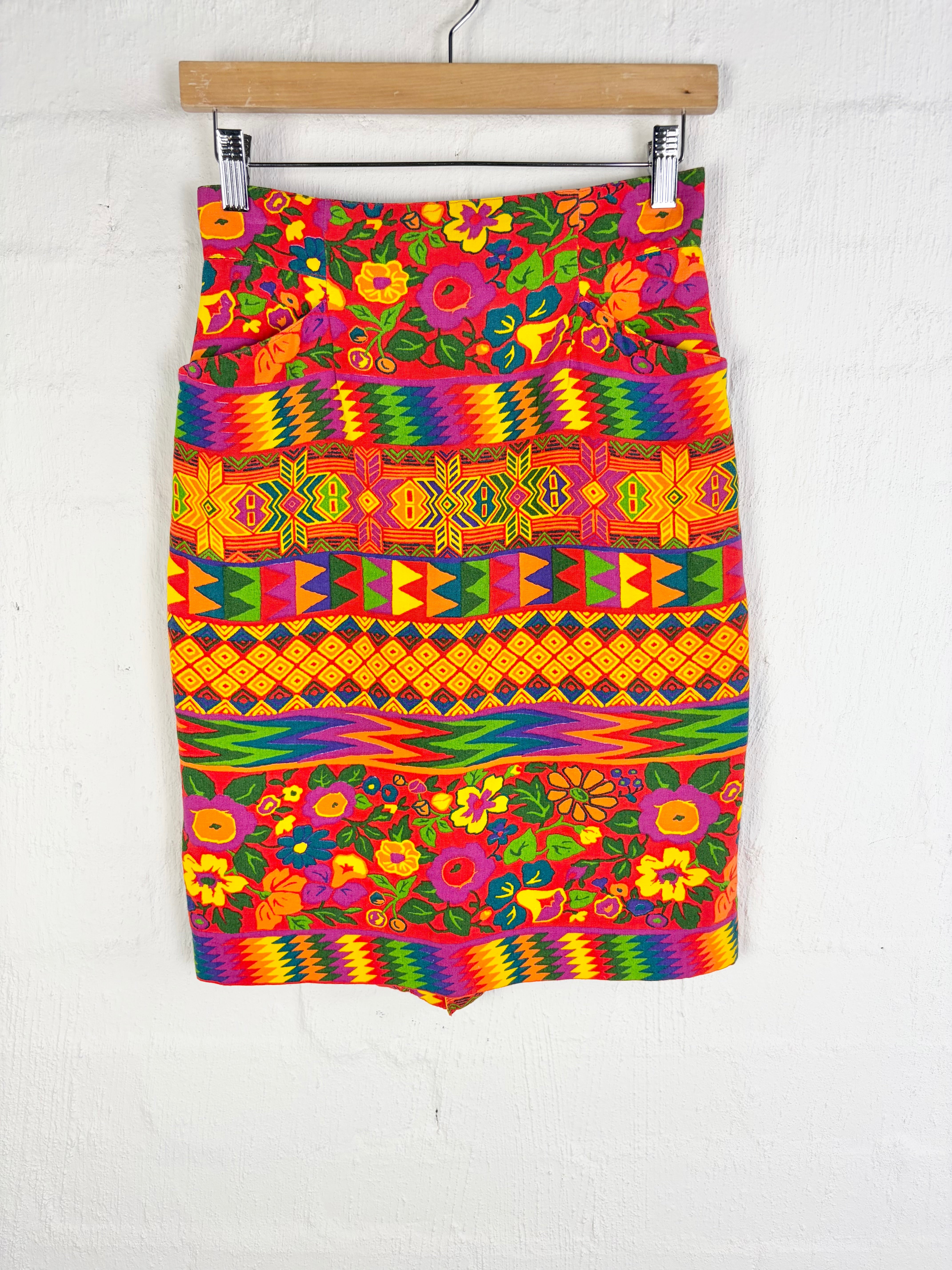 A vibrant, multicolored skirt with intricate floral and geometric patterns, hanging from a wooden hanger against a white textured wall.