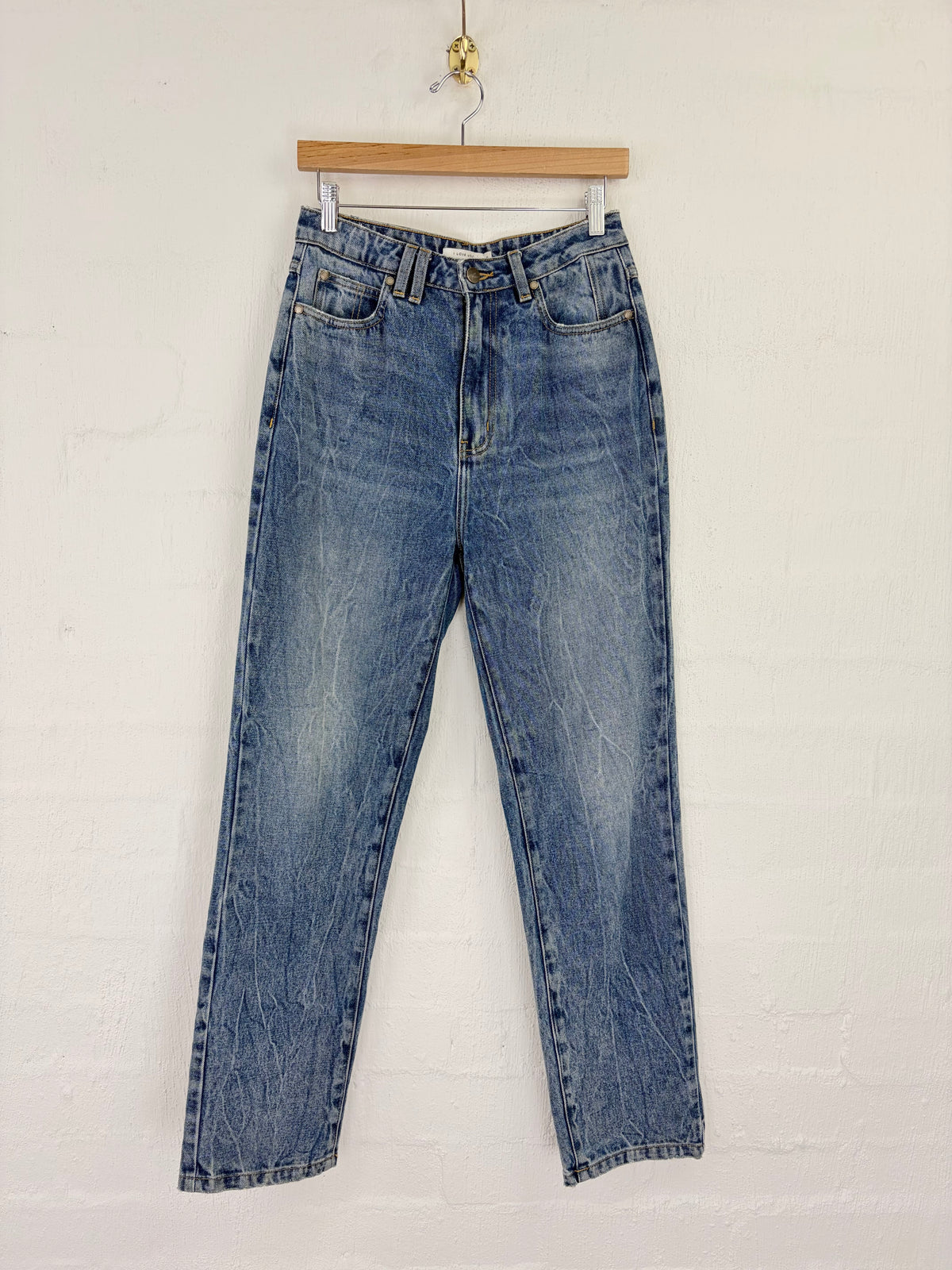 A pair of high-waisted, straight-leg blue denim jeans with faded whiskering and whiskering details, hanging from a wooden hanger against a white painted brick wall.