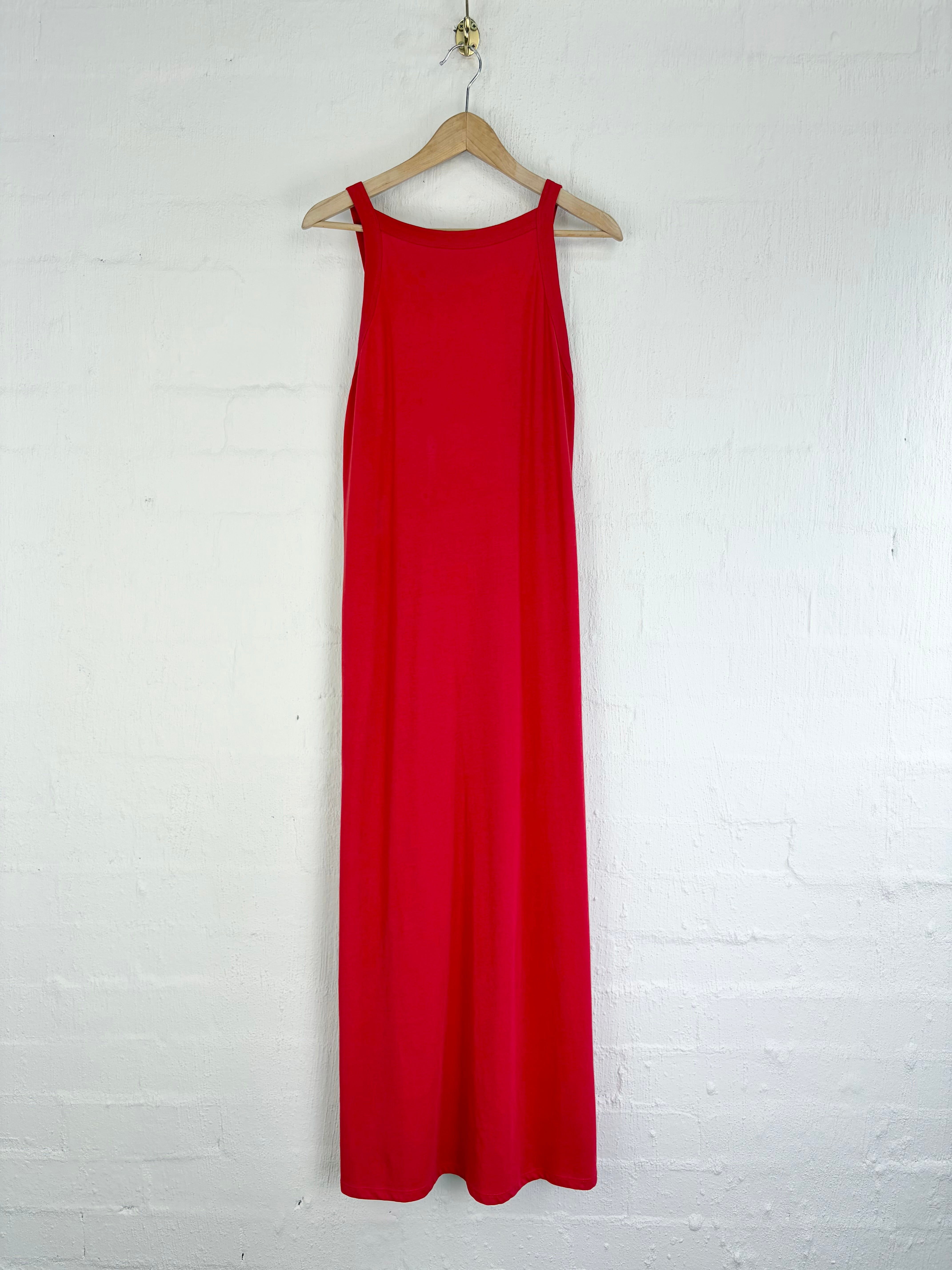 A bright red sleeveless maxi dress hanging on a wooden hanger against a white painted brick wall.