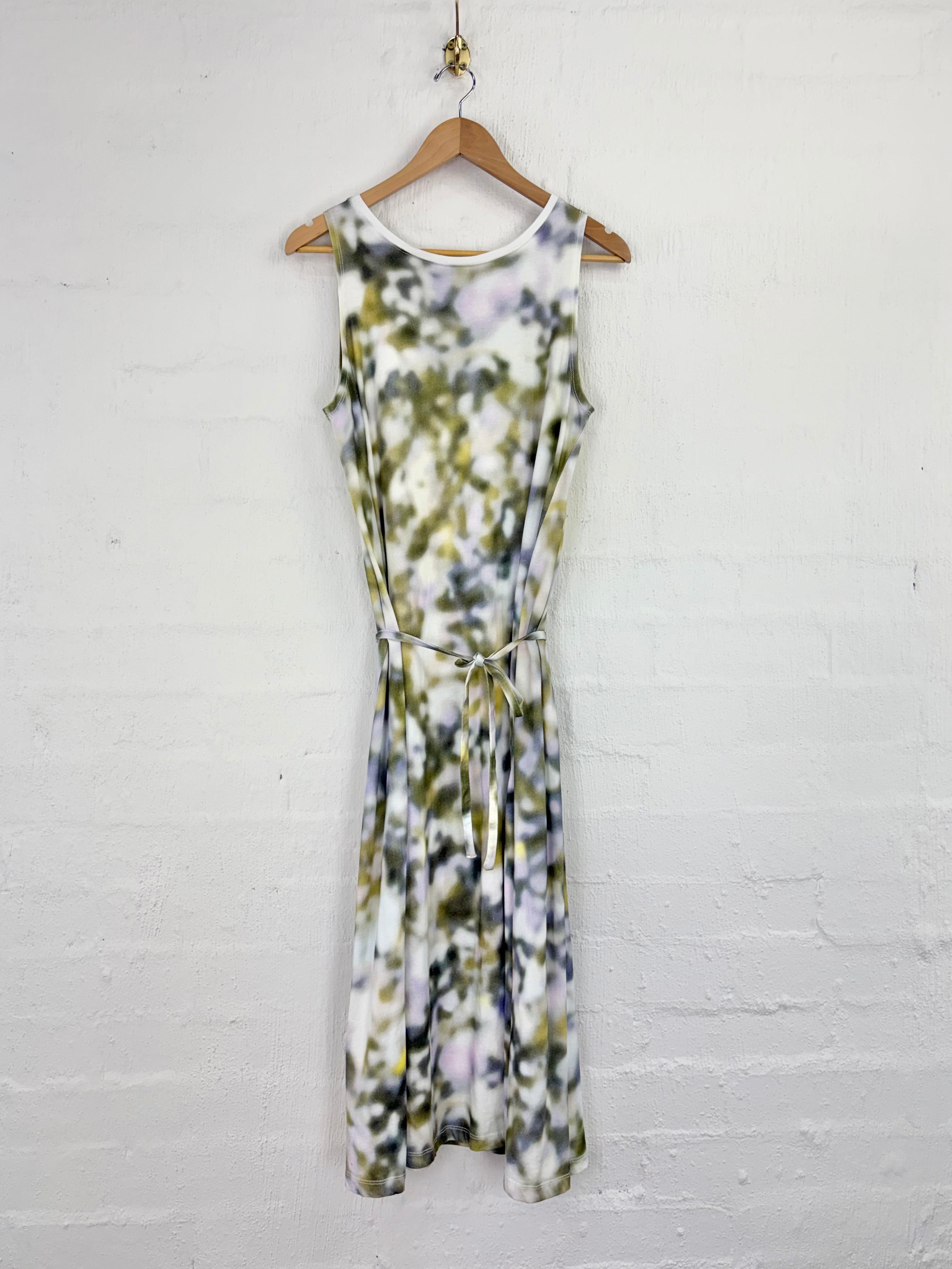 A sleeveless midi dress with a tie-dye pattern in olive green, purple, and white, hanging on a wooden hanger against a white painted brick wall.