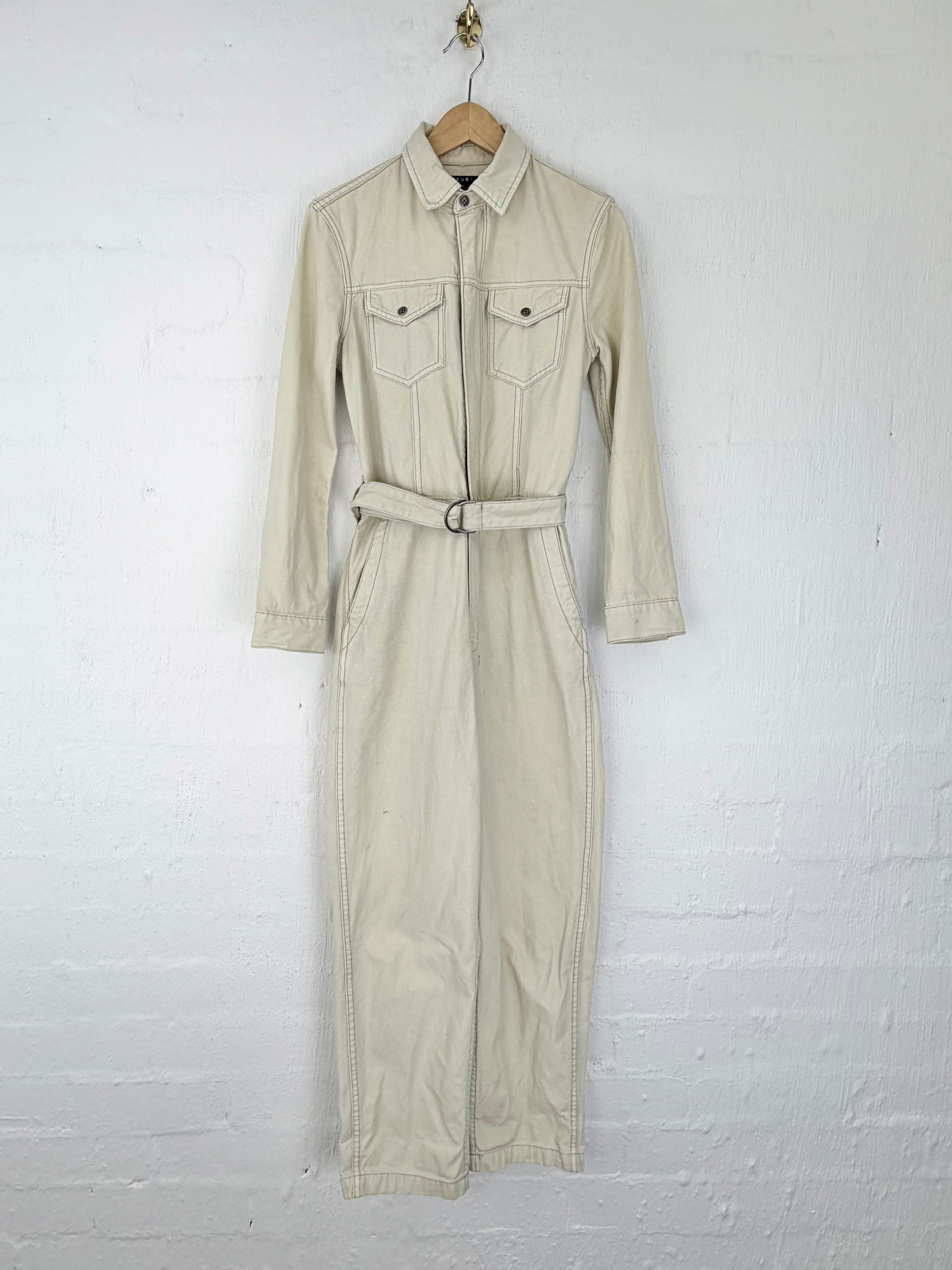 A light beige utility jumpsuit with a matching waist belt, hanging on a wooden hanger against a white painted brick wall.