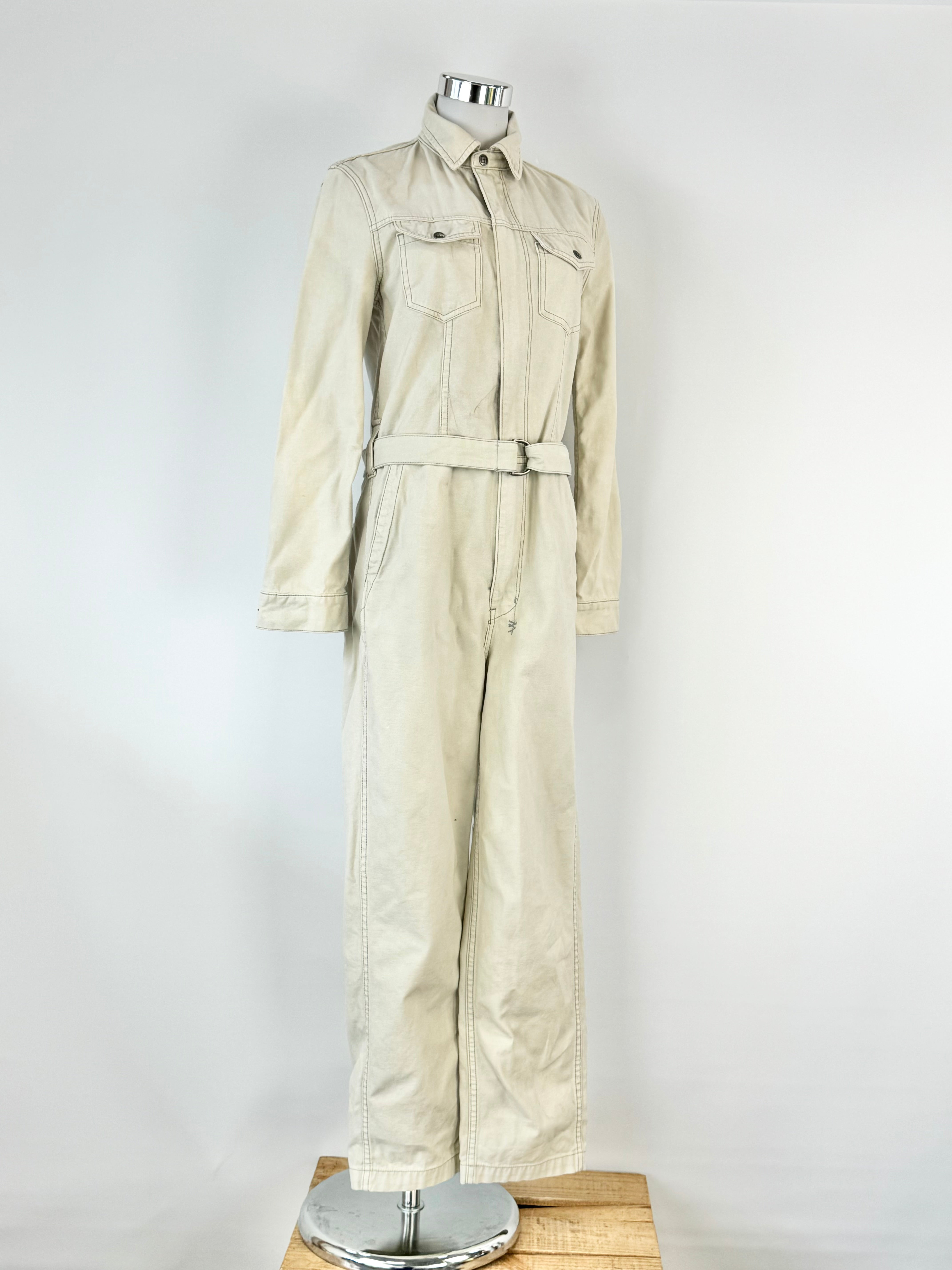 A long sleeve beige cotton jumpsuit with multiple pockets and a button-up front, displayed on a mannequin.