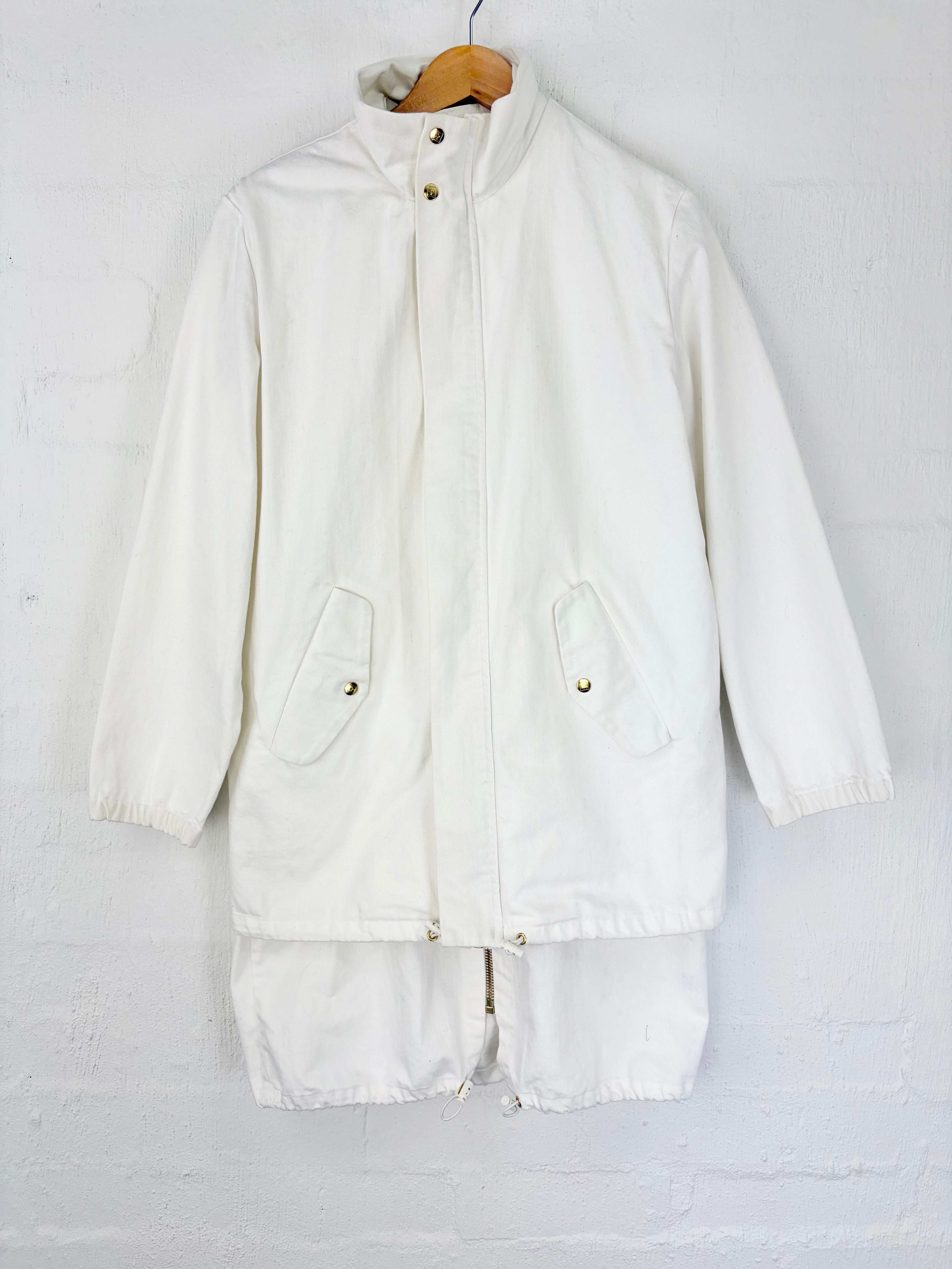 An off-white canvas utility jacket with a stand-up collar, front snap closure, and two front flap pockets, hanging on a wooden hanger against a white painted brick wall.