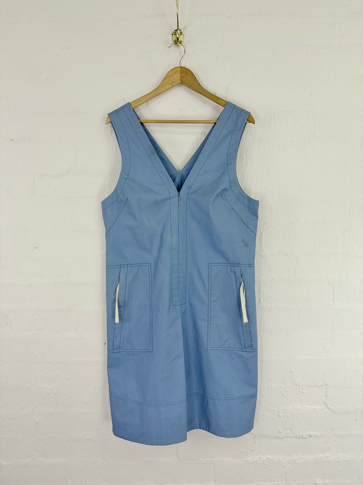 A light blue sleeveless utility vest with a V-neck and front pockets, hanging on a wooden hanger against a white brick wall.