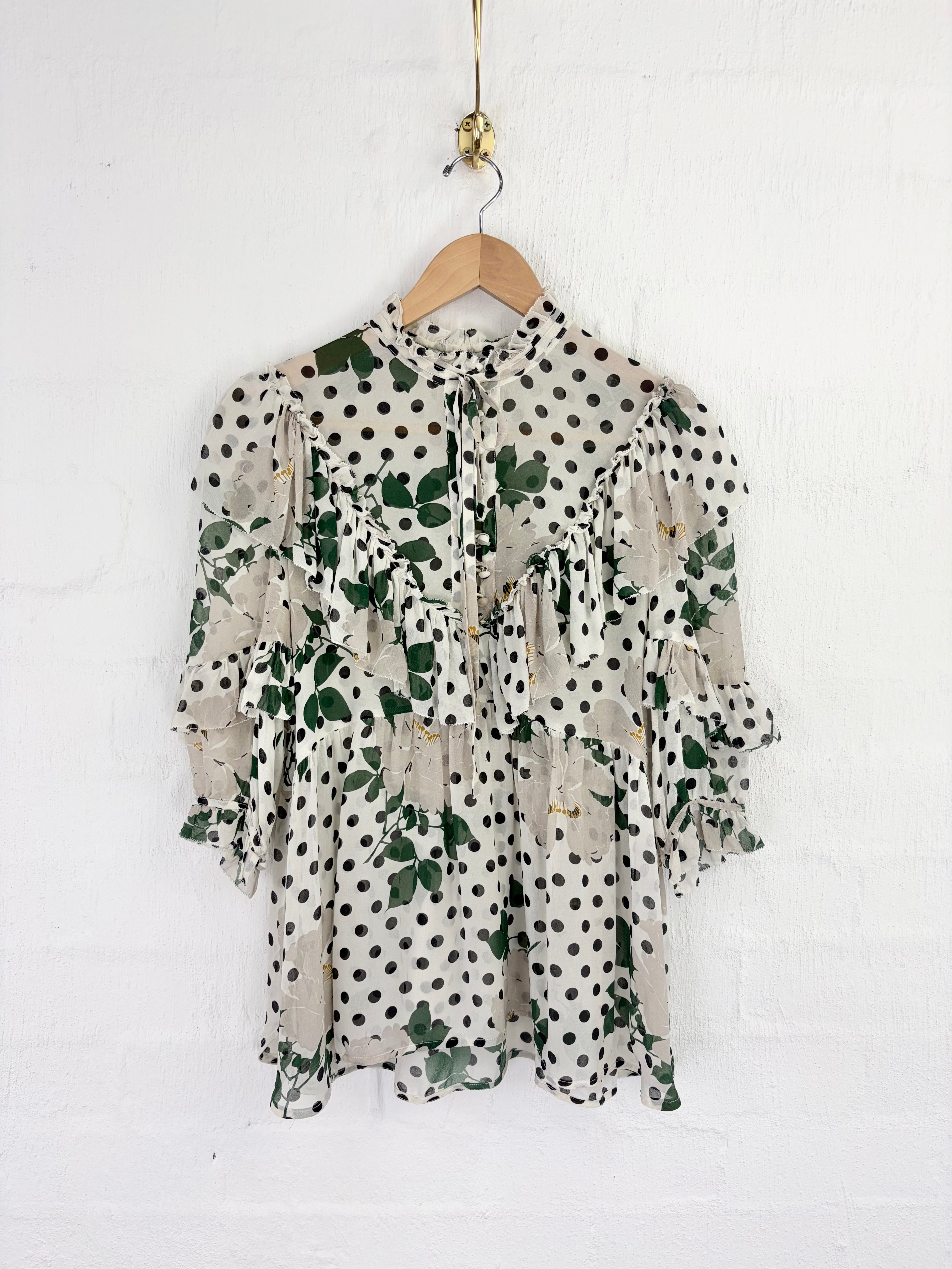 A ruffled long-sleeve blouse with a green and black polka dot floral print, hanging on a wooden hanger against a white painted brick wall.