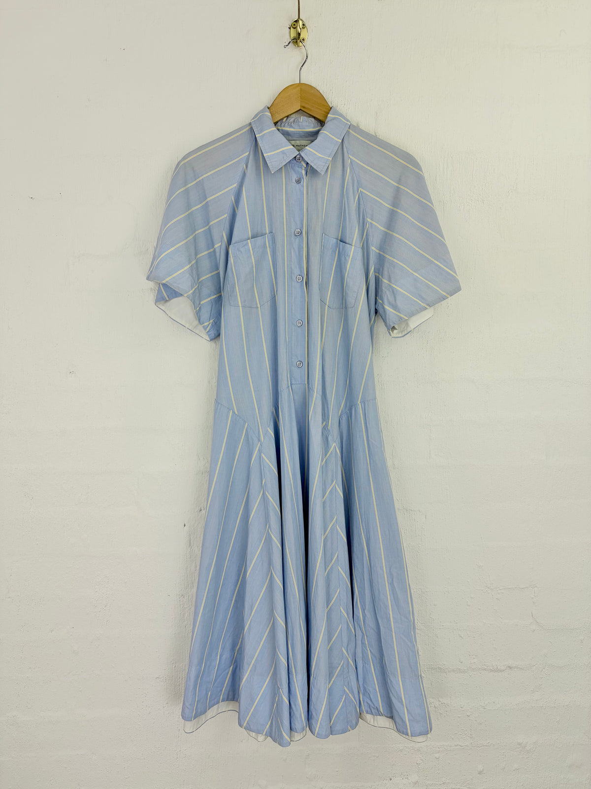 A light blue short-sleeve button-up shirt dress with thin white vertical stripes, hanging on a wooden hanger against a white painted brick wall.