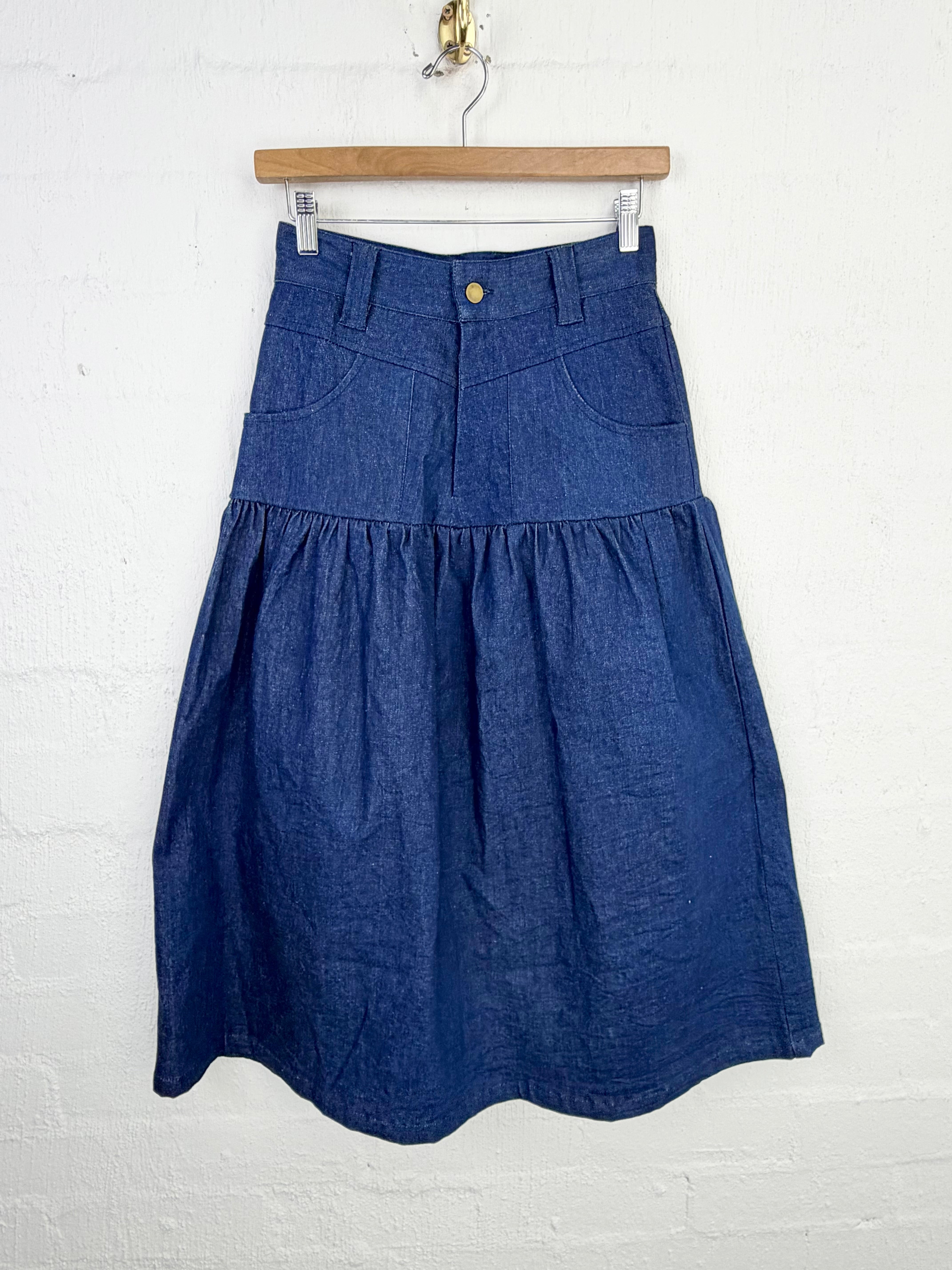 A blue denim tiered skirt with a front button closure and side pockets, hanging from a wooden hanger against a white painted brick wall.