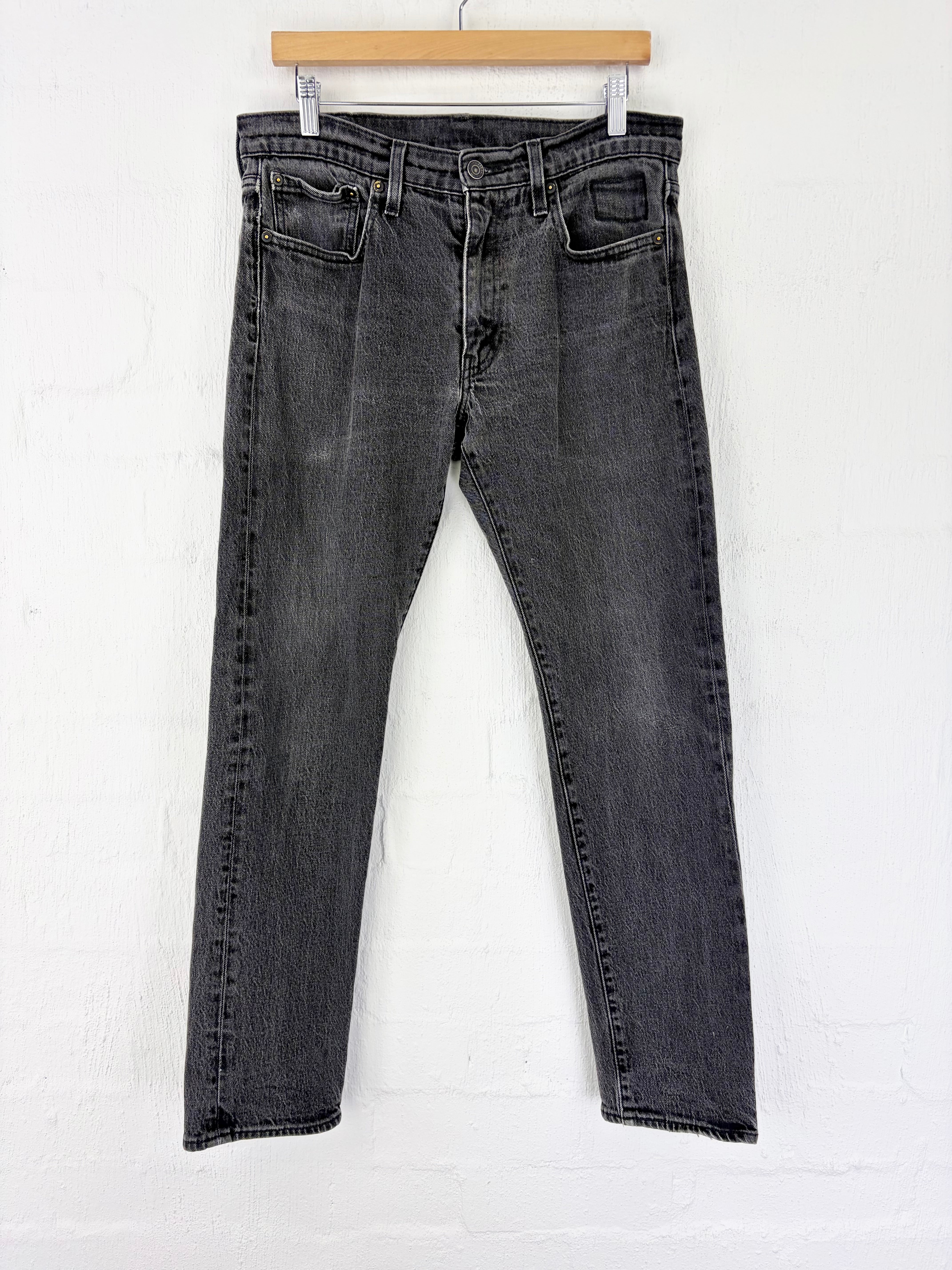 A pair of dark gray washed denim jeans hanging from a wooden hanger against a white brick wall.
