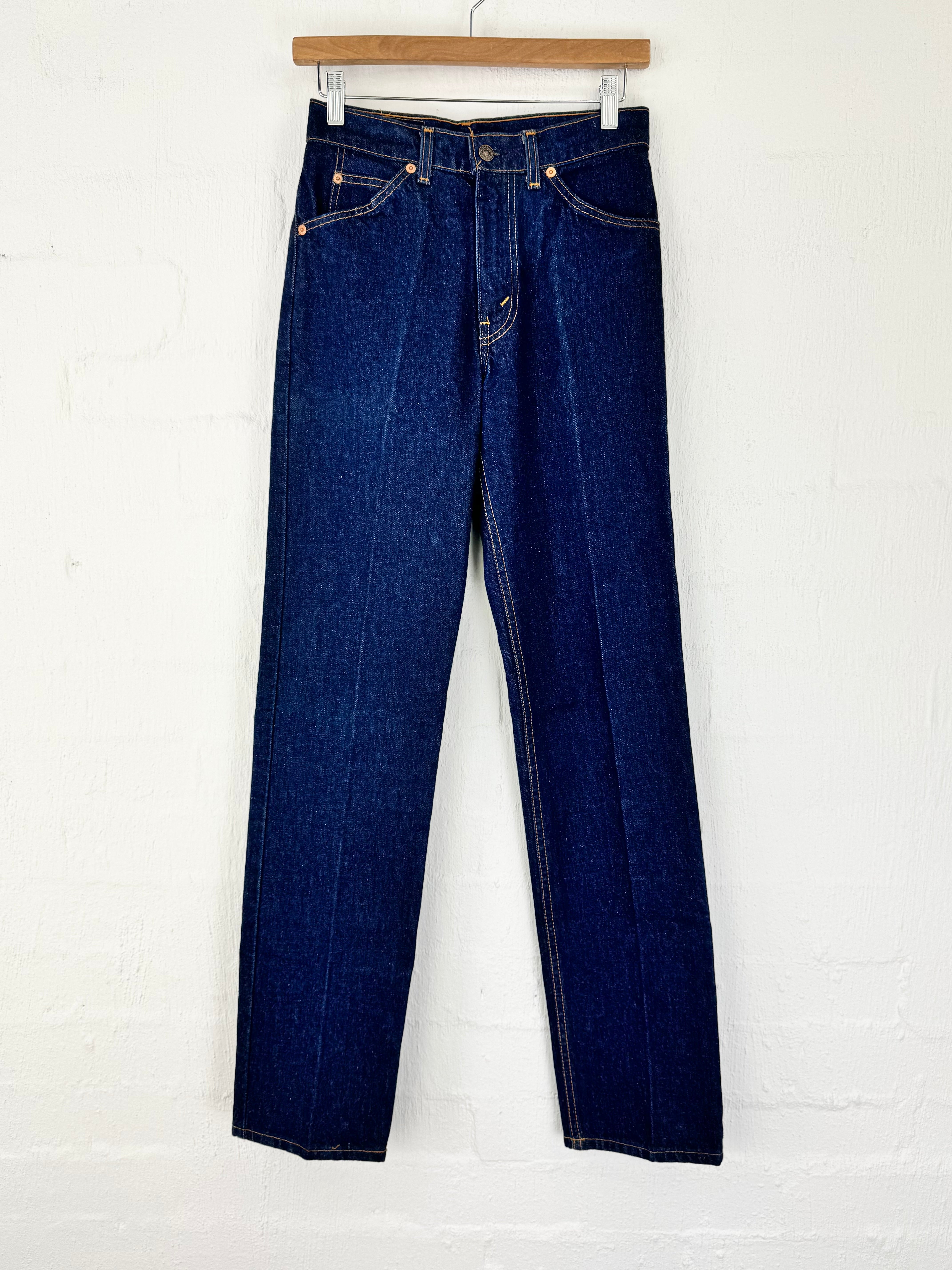 A pair of dark blue denim jeans hanging from a wooden hanger against a white painted brick wall.