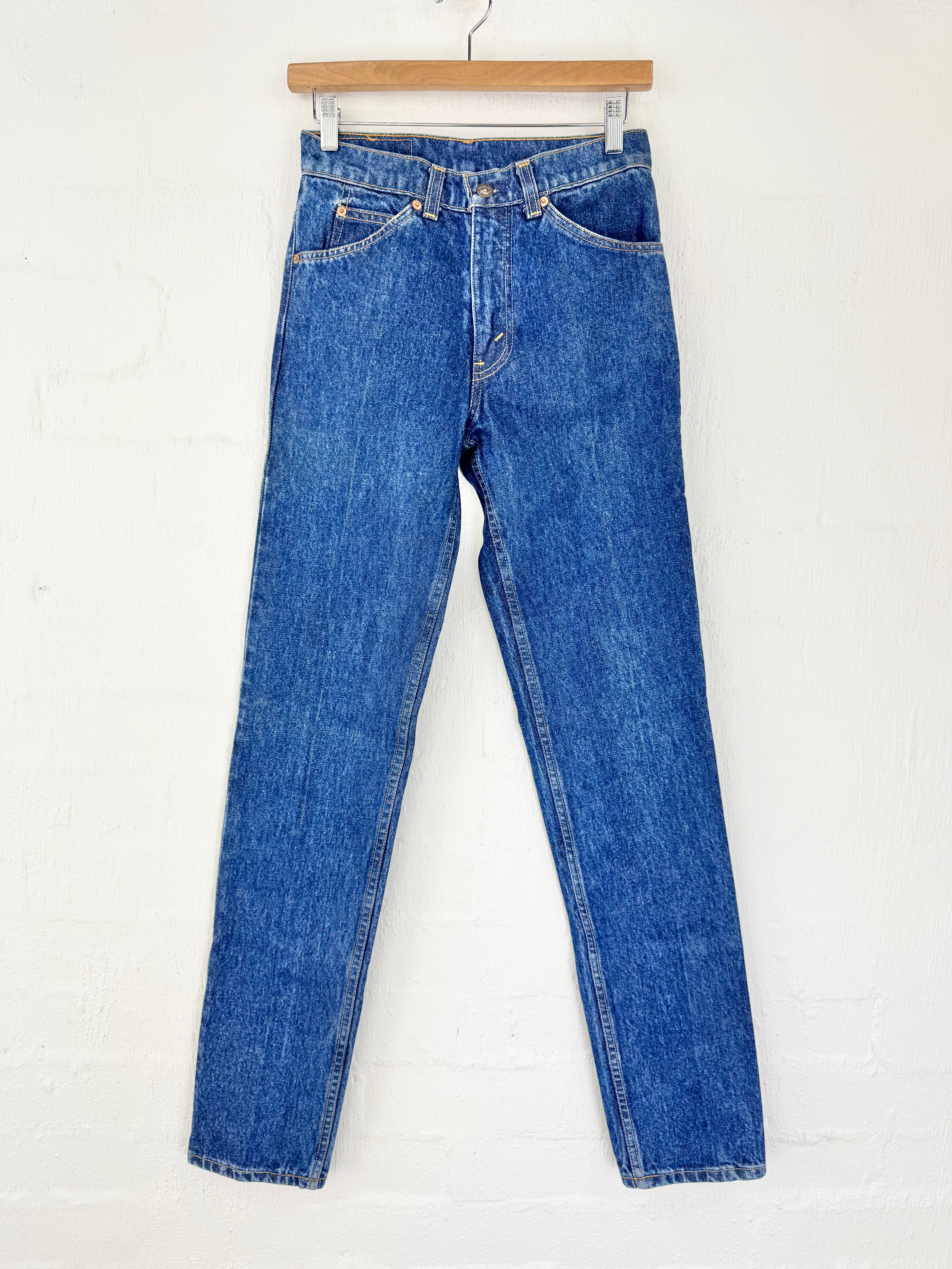 A pair of medium-wash blue denim jeans hanging from a wooden hanger against a white painted brick wall.