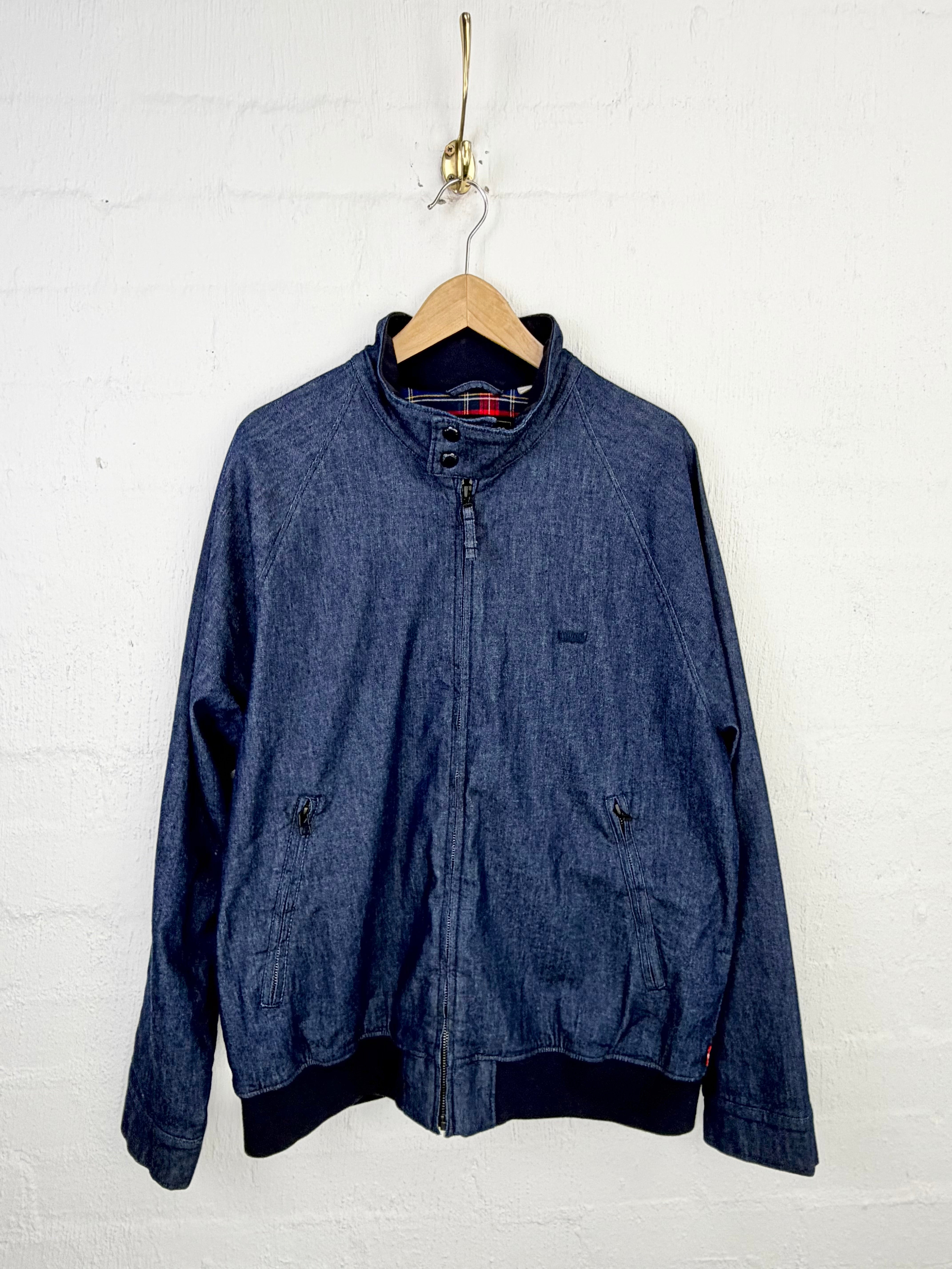A blue denim zip-up jacket hanging on a wooden hanger against a white painted brick wall.