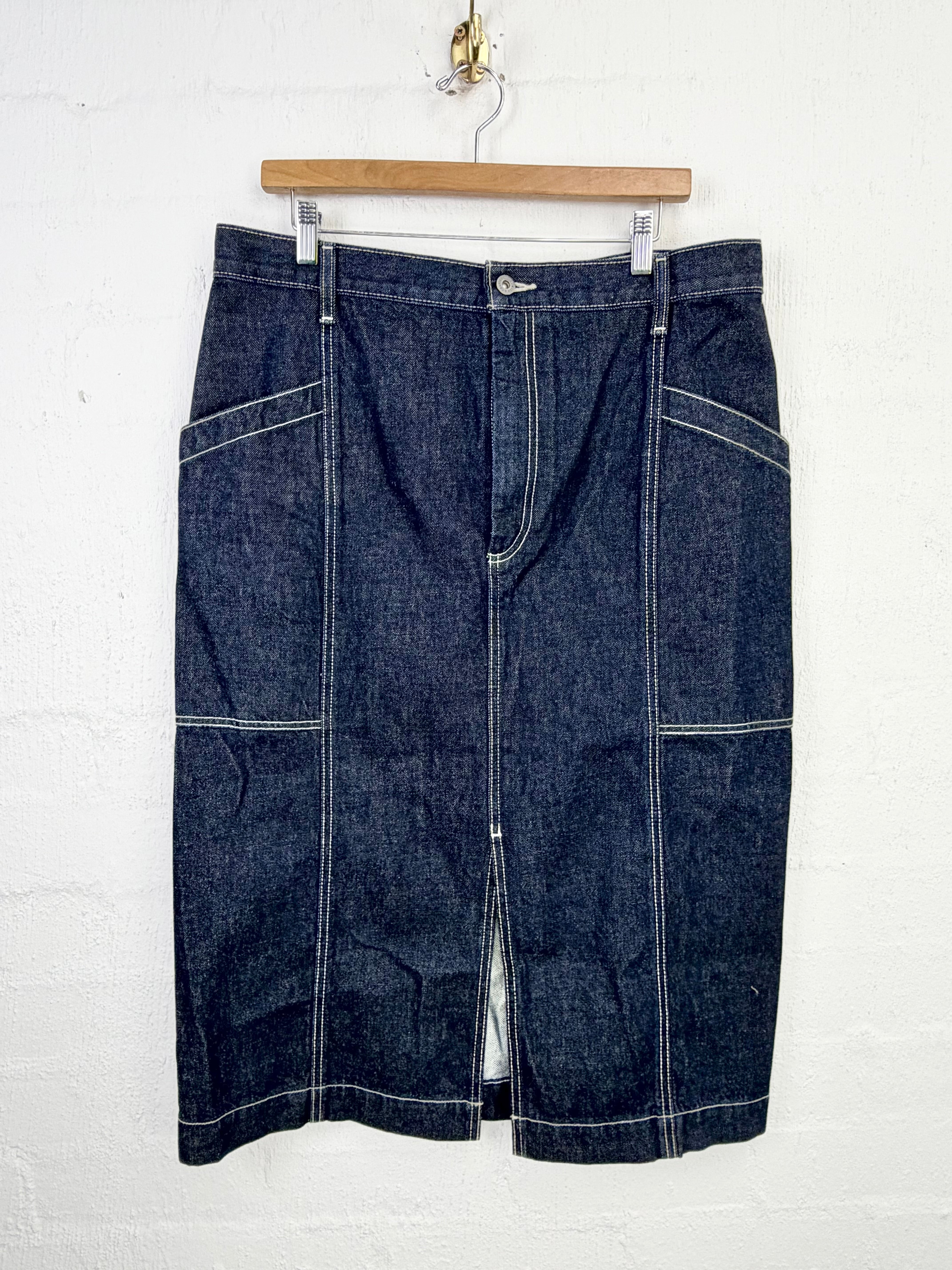 A dark indigo denim skirt with contrast white stitching, hanging from a wooden hanger against a white painted brick wall.