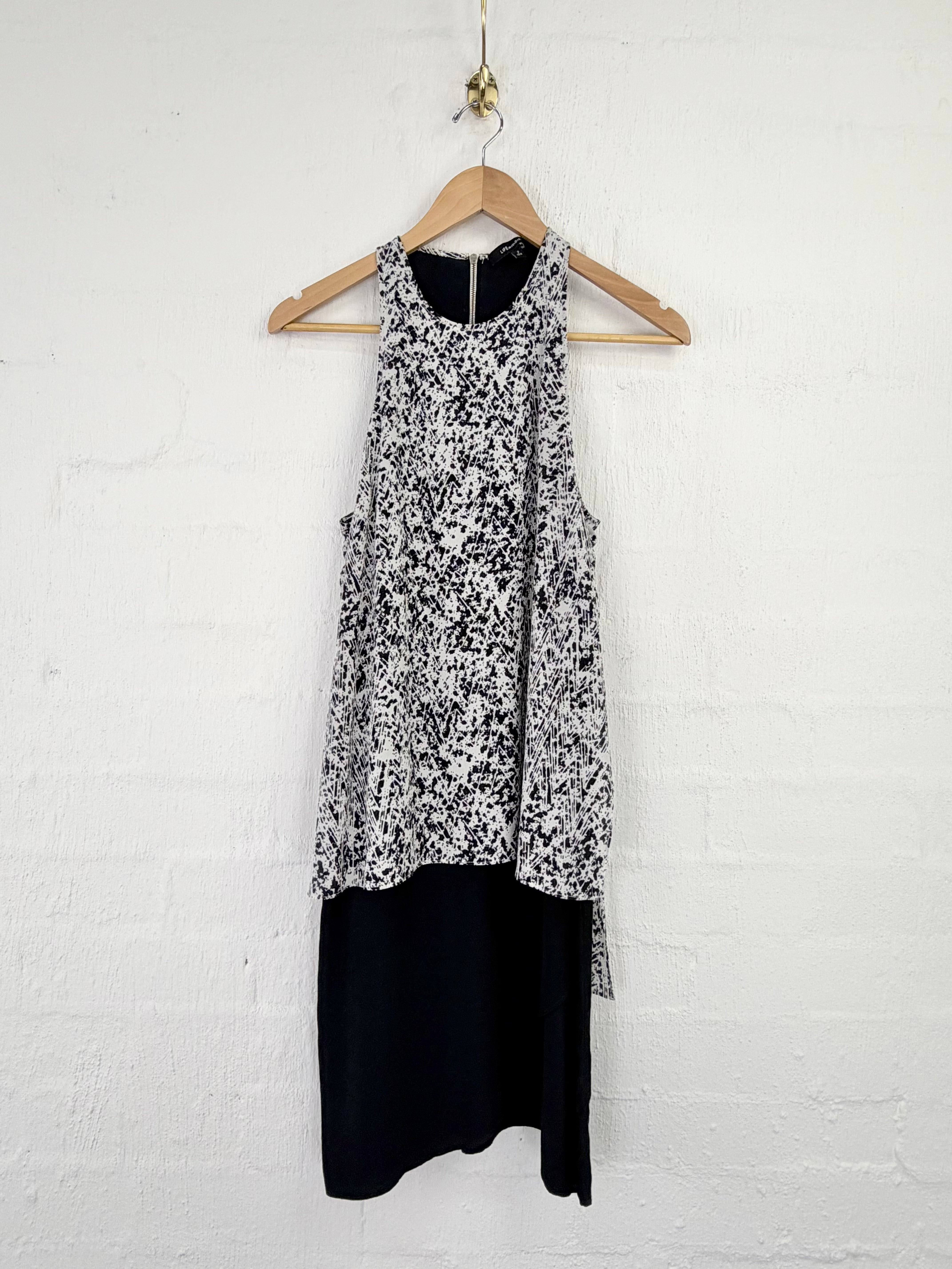 A sleeveless black and white patterned top layered over a solid black skirt, hanging on a wooden hanger against a white painted brick wall.