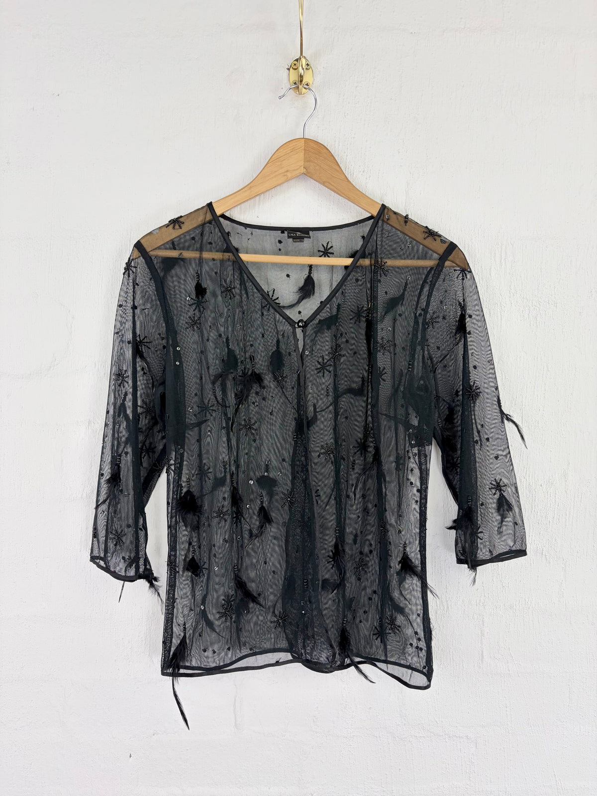 A sheer black mesh blouse with black feather embellishments, hanging on a wooden hanger against a white painted brick wall.