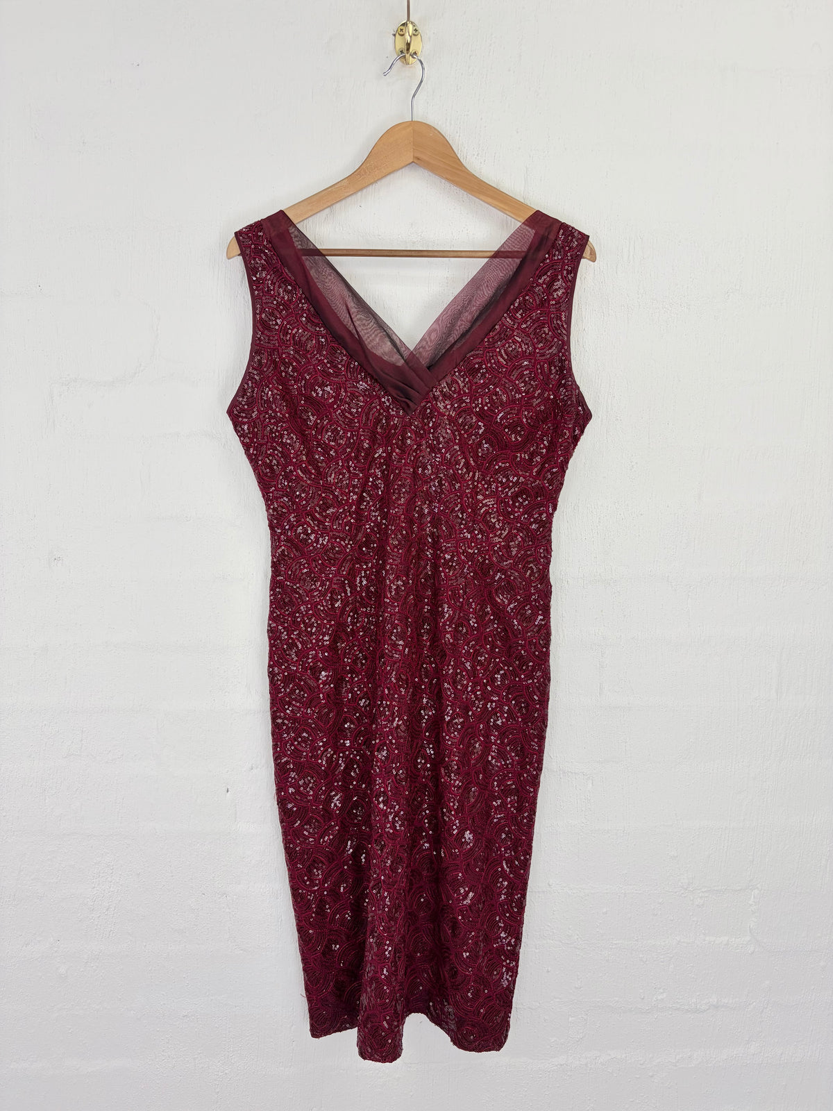 A sleeveless burgundy lace dress with a sheer V-neckline, hanging on a wooden hanger against a white painted brick wall.