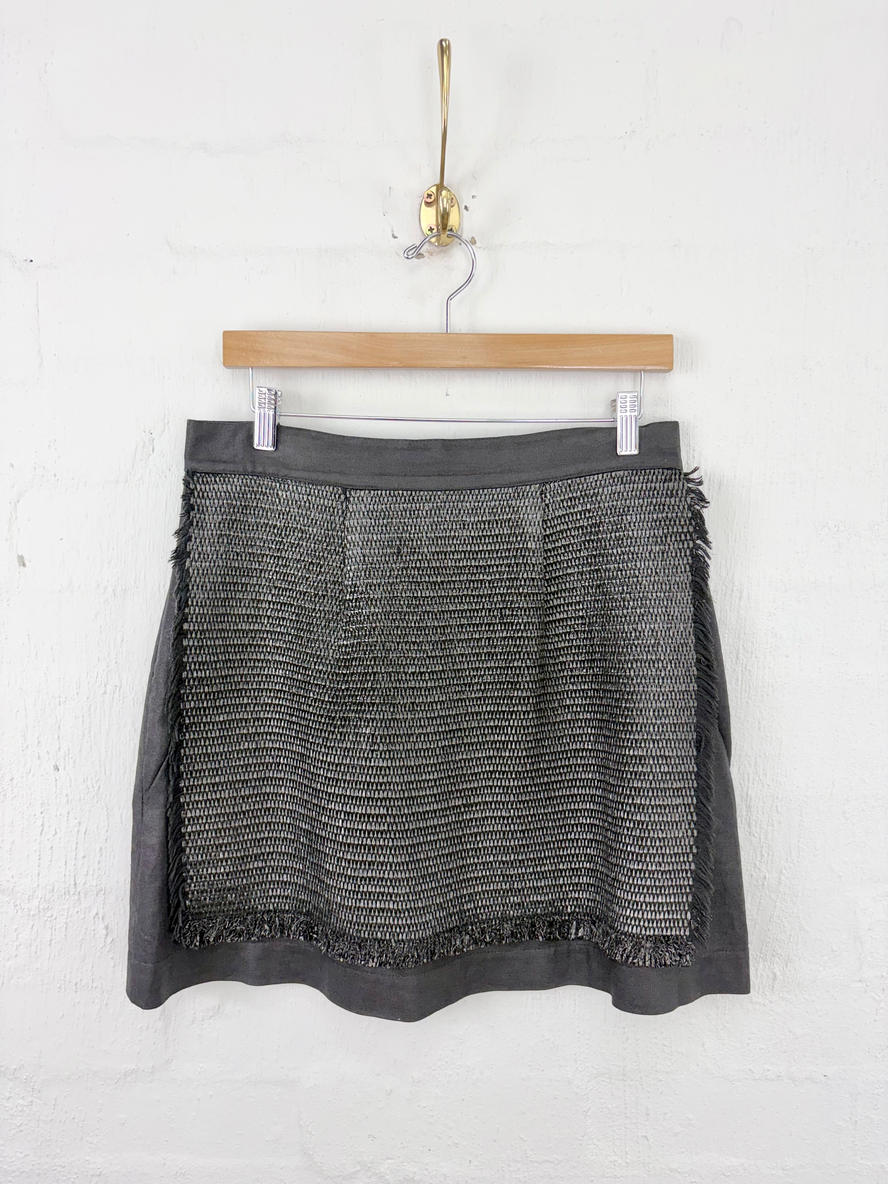 A dark gray textured tweed mini skirt with fringed edges, hanging on a wooden hanger against a white brick wall.