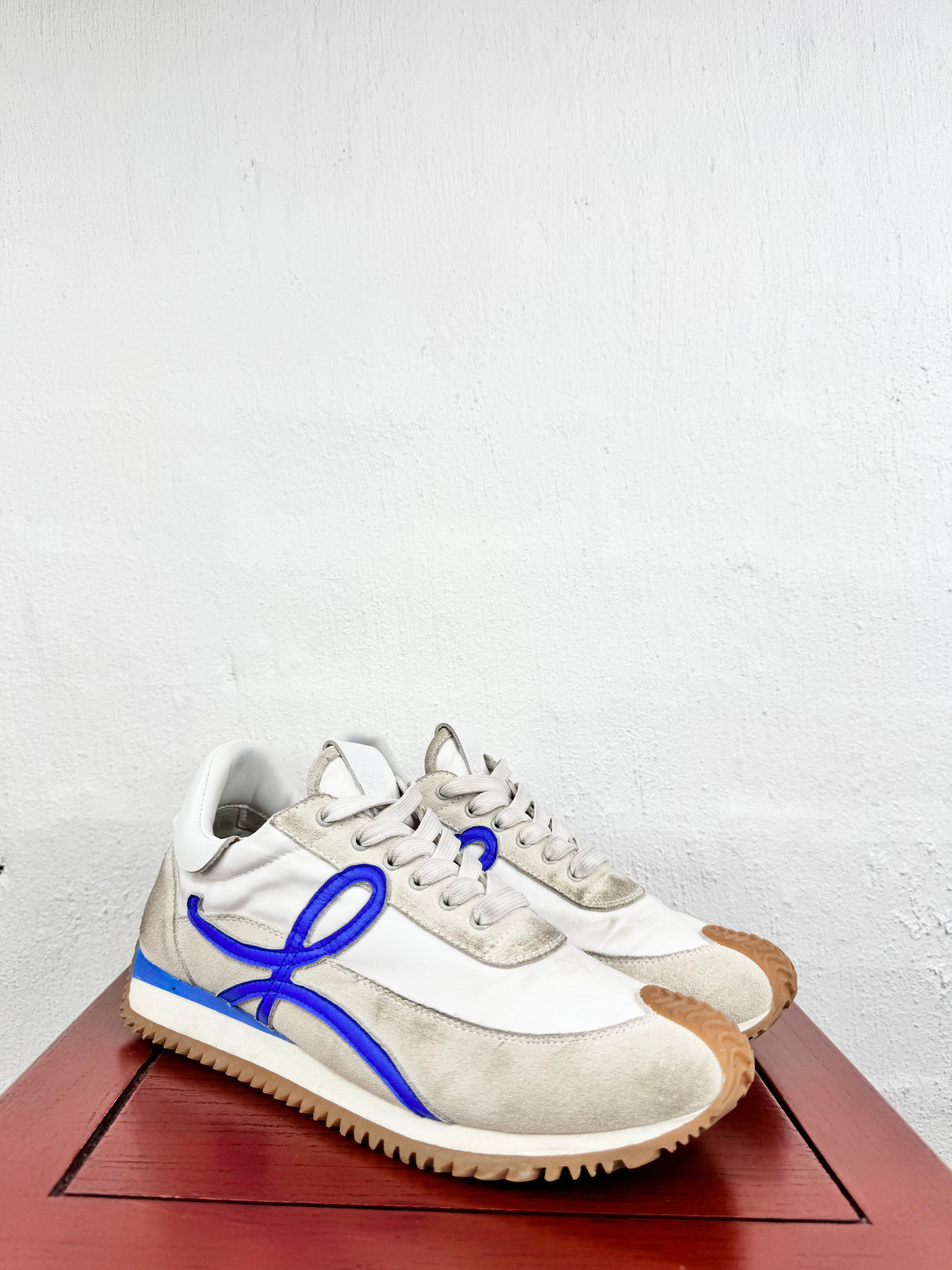 A pair of vintage Onitsuka Tiger sneakers in off-white suede and leather, featuring a vibrant blue bow detail on the side and a brown gum sole, displayed on a reddish-brown wooden surface against a white wall.