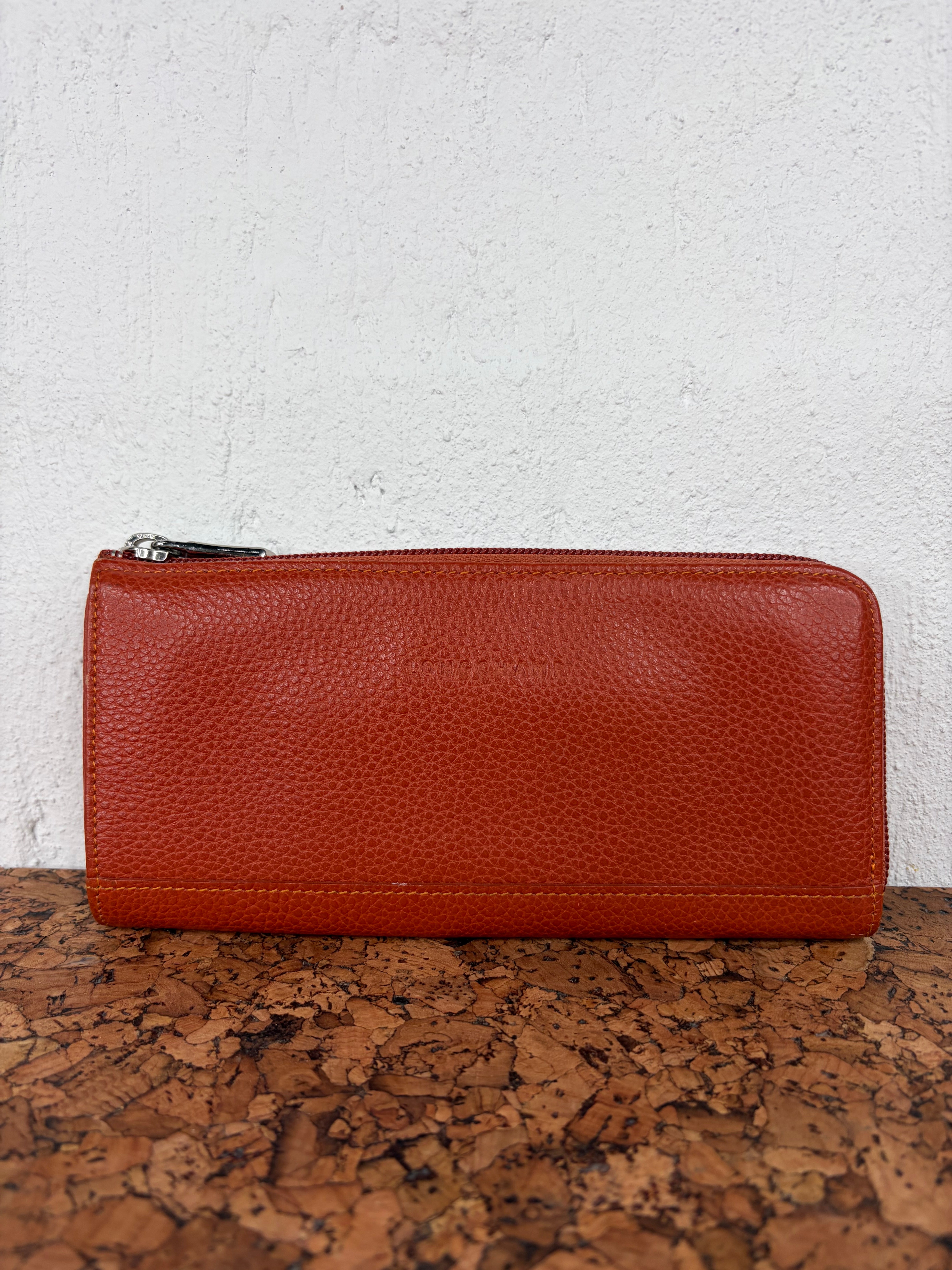 A reddish-brown pebbled leather zip-around wallet with a silver zipper pull, resting on a cork surface against a white textured wall.