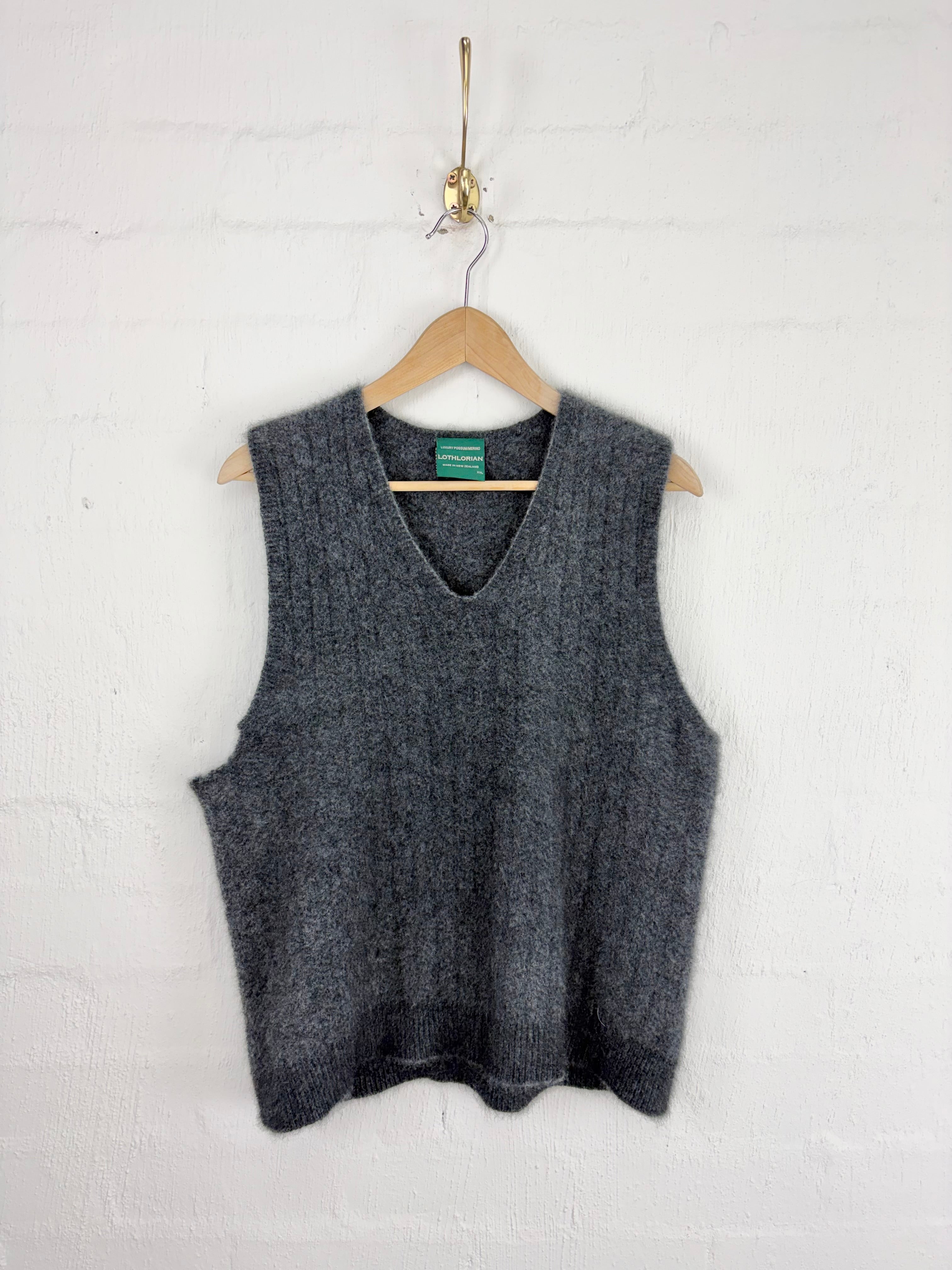 A dark gray ribbed knit sweater vest hanging on a wooden hanger against a white painted brick wall.