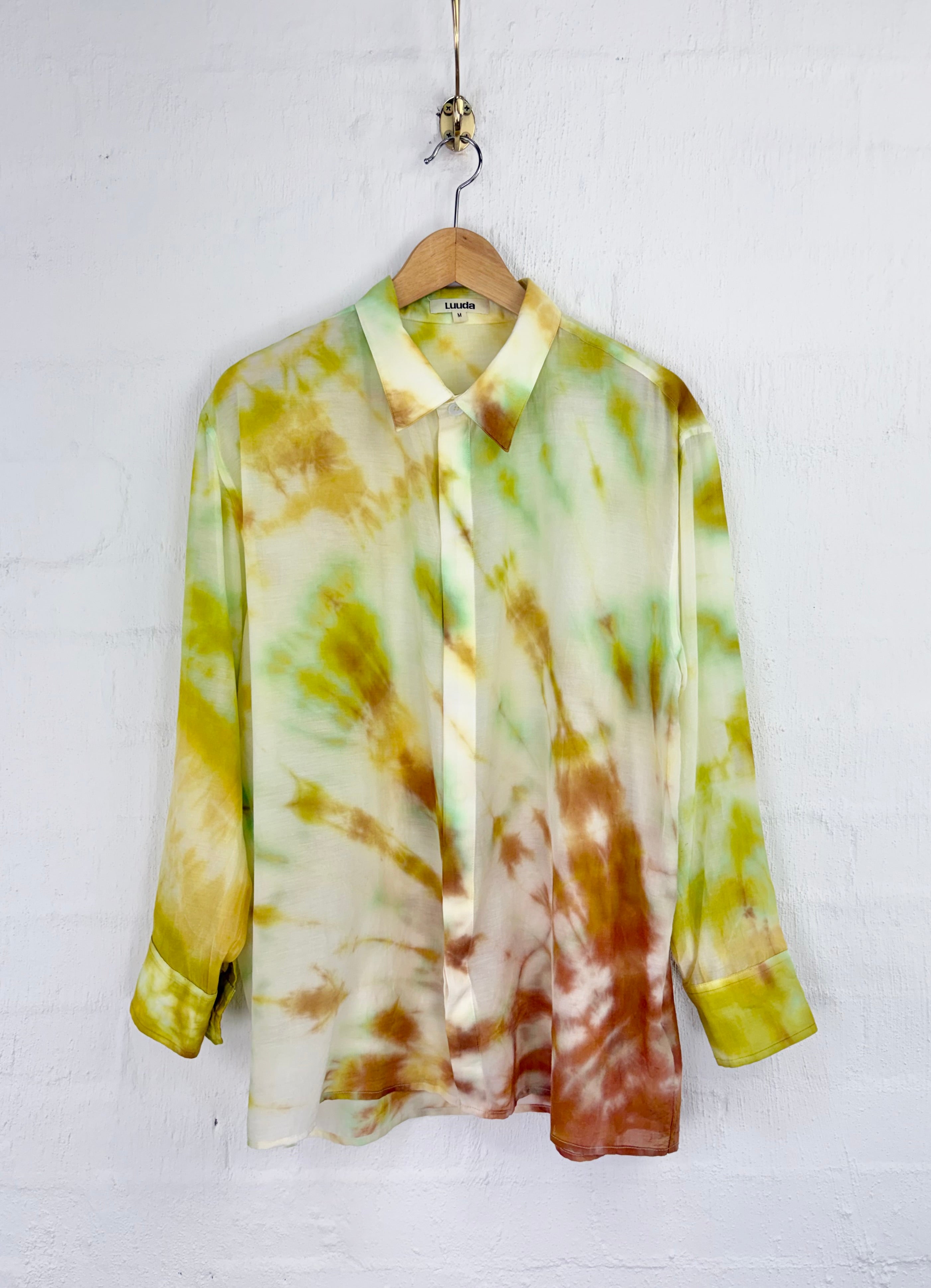 A long-sleeve collared blouse with a tie-dye pattern in olive green, mustard yellow, and rust orange, hanging on a wooden hanger against a white painted brick wall.