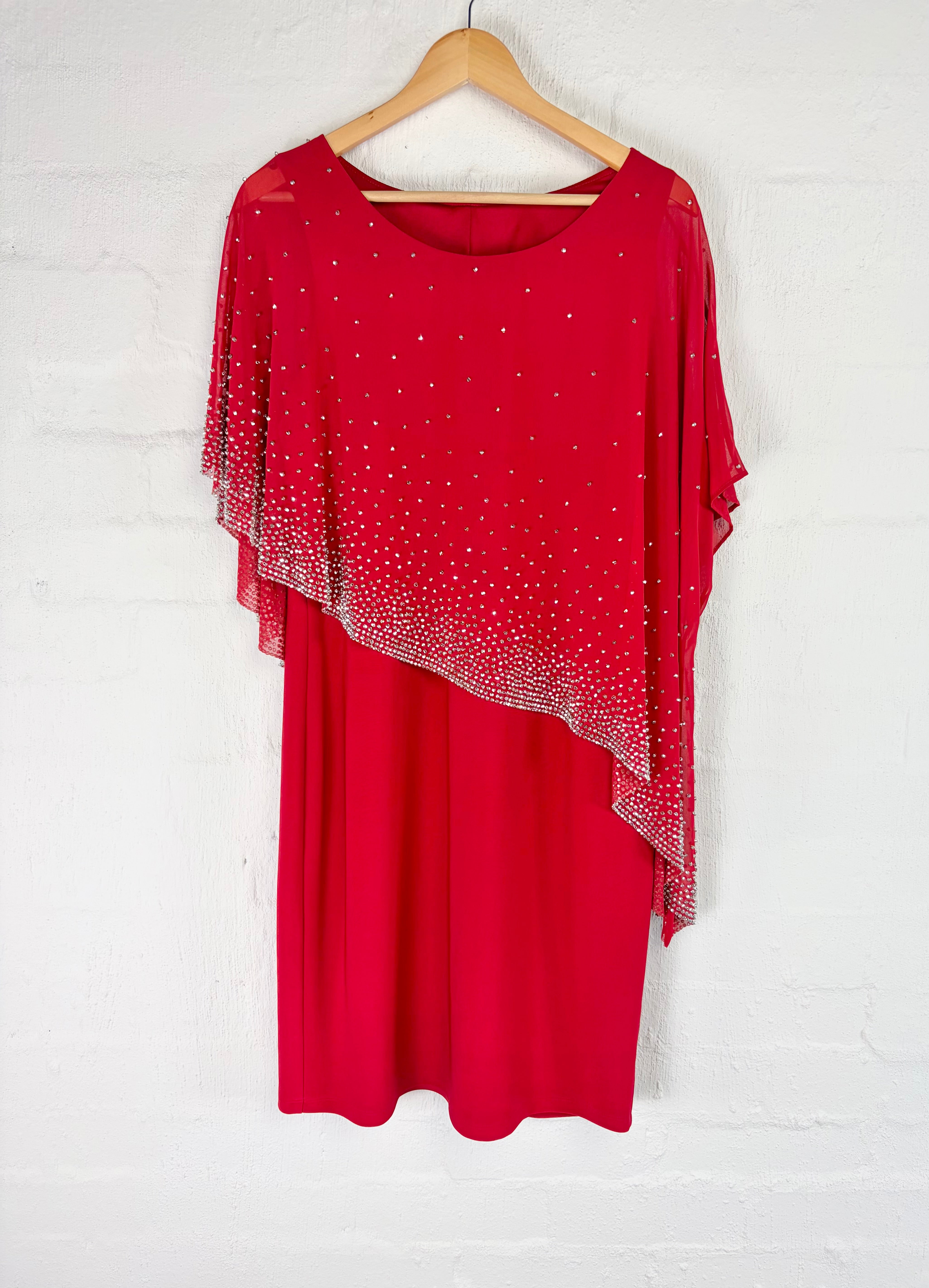 A red short-sleeve dress with a sheer, embellished overlay draped over the shoulders, hanging on a wooden hanger against a white brick wall.