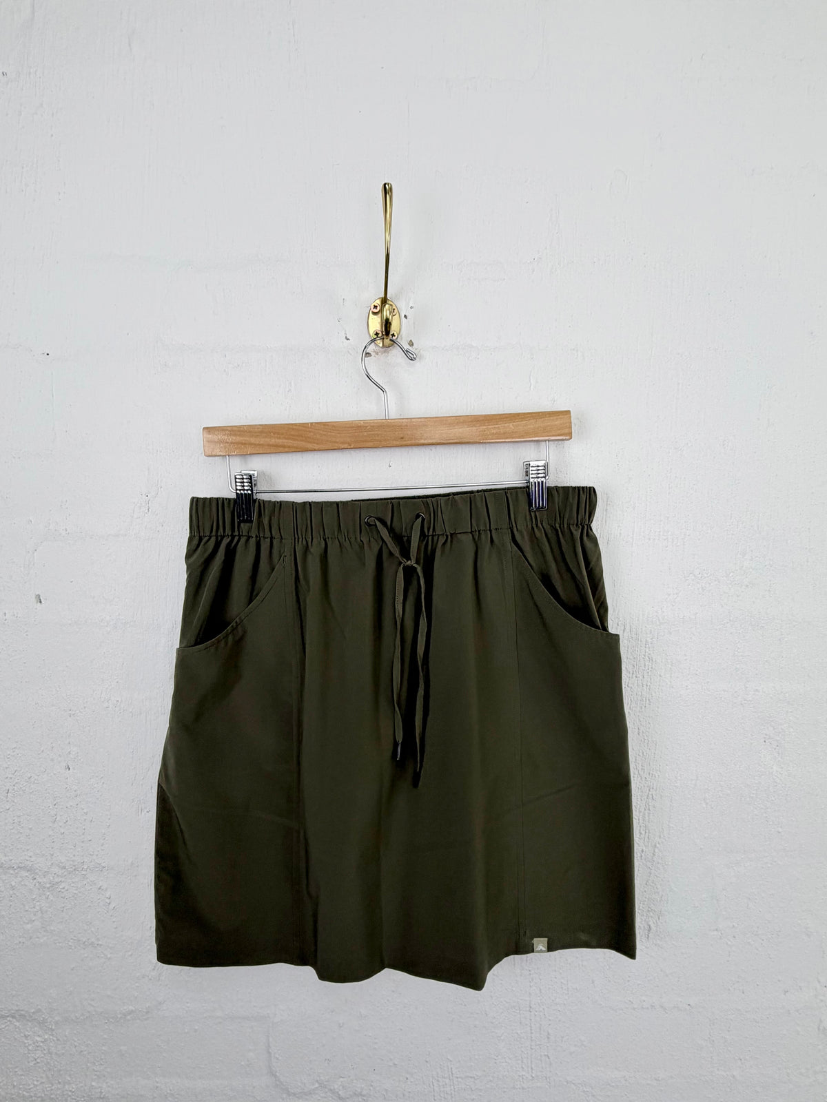 Olive green drawstring shorts with side pockets, hanging from a wooden hanger on a white painted brick wall.