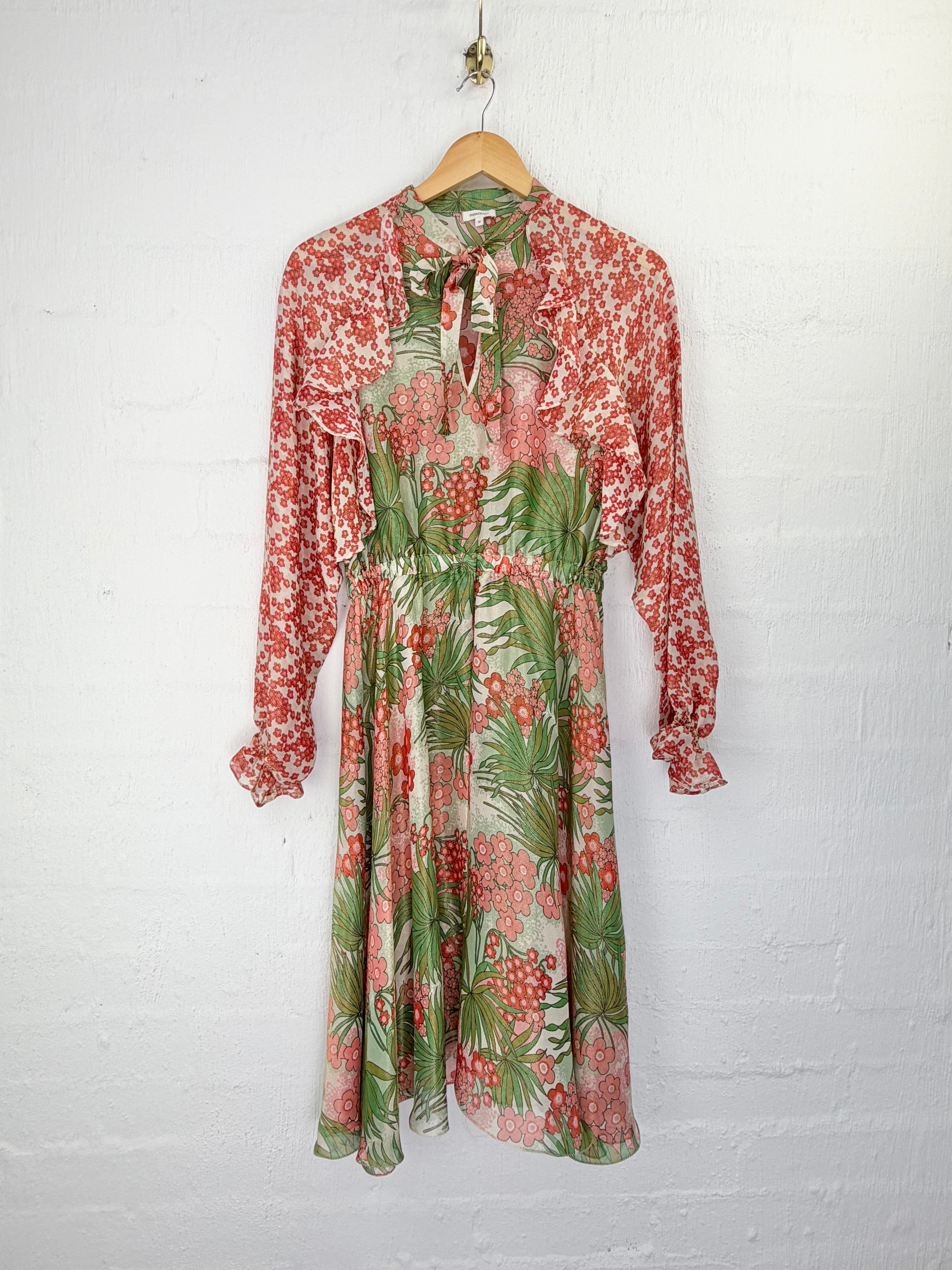 A long-sleeve midi dress with a tie-neck and ruffled shoulders, featuring a tropical floral print in red, green, and beige, hanging on a wooden hanger against a white brick wall.