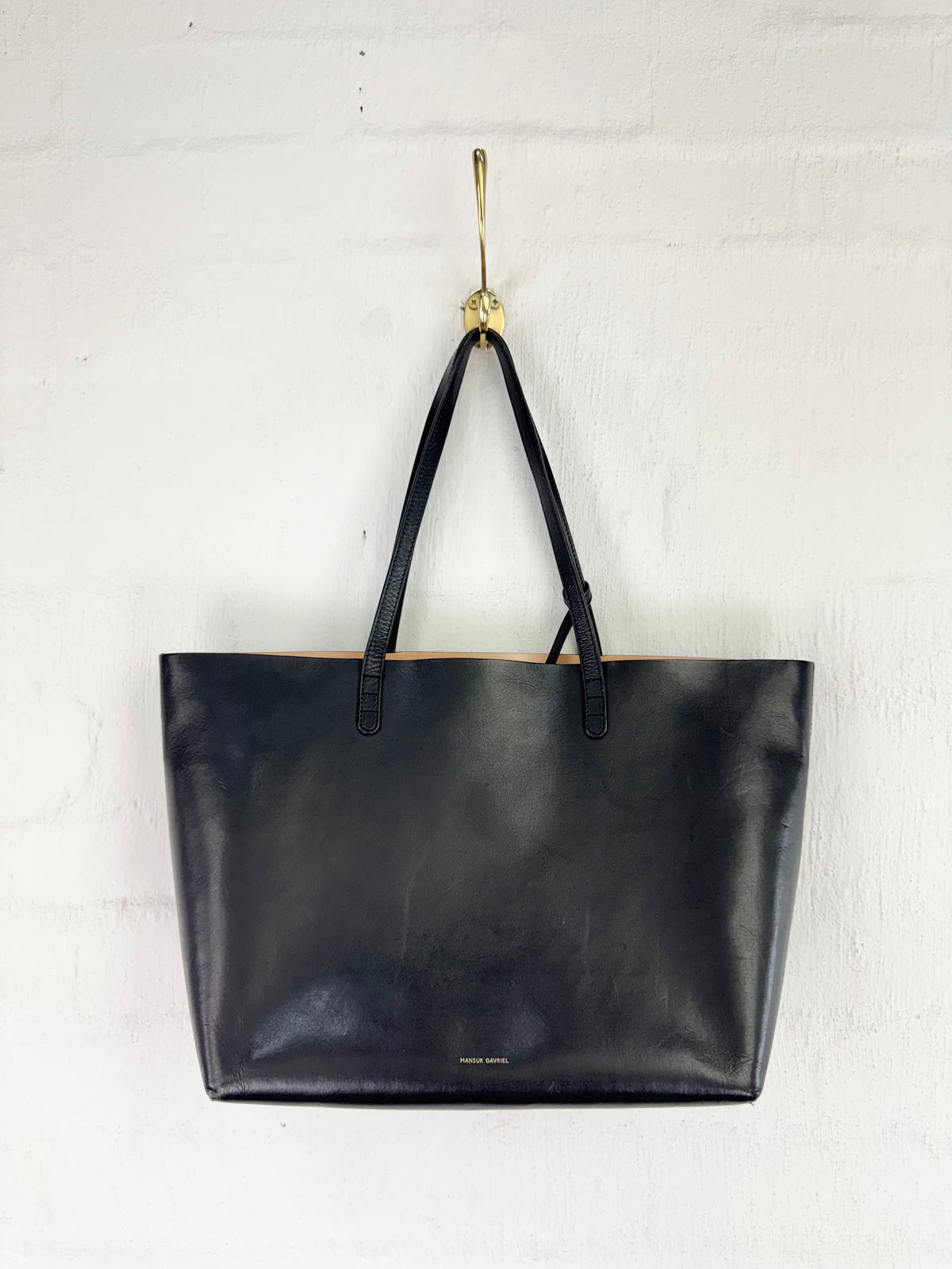 A black leather tote bag with double shoulder straps, hanging from a gold hook on a white painted brick wall.
